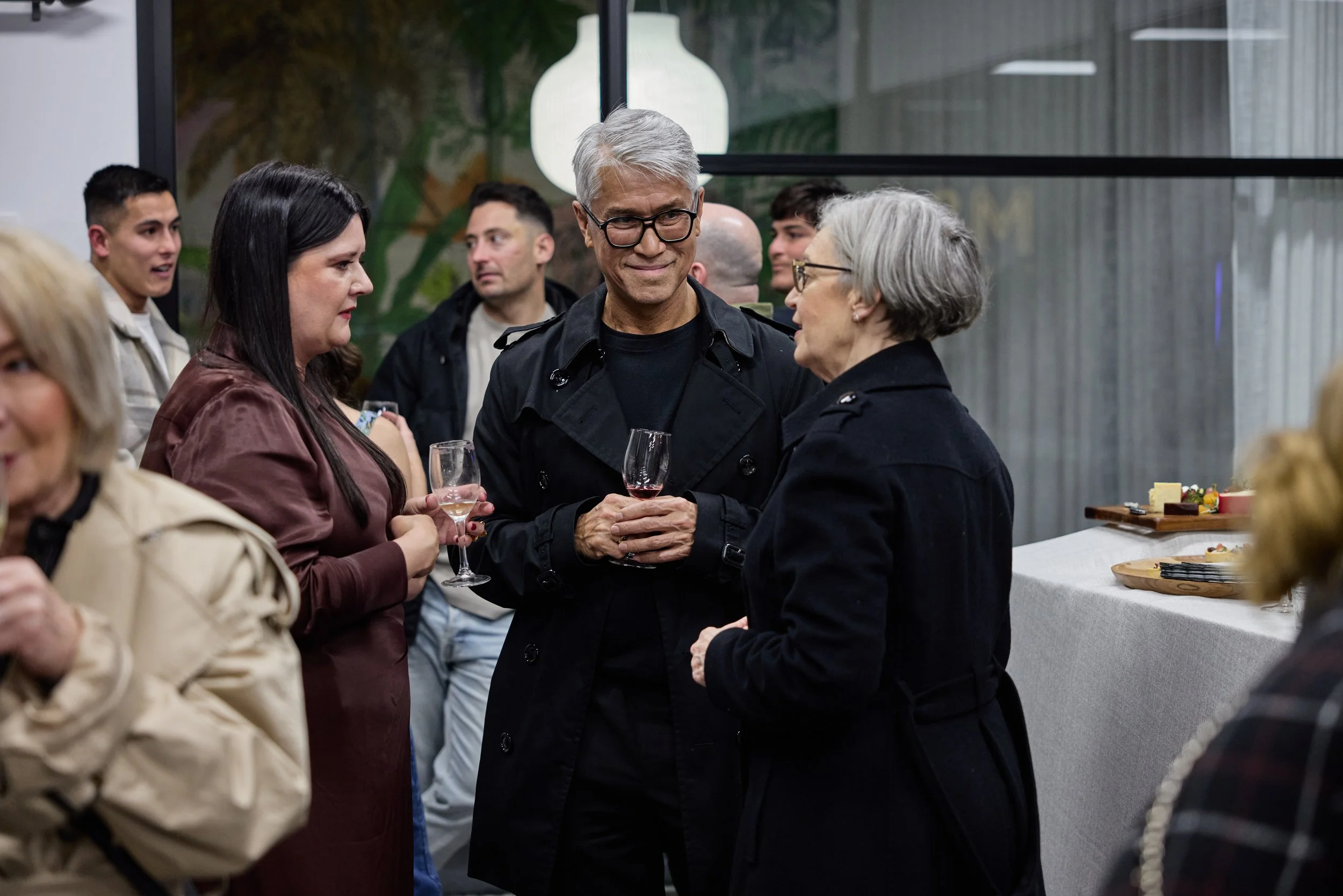 People socializing at a gathering, with a woman in a brown jacket holding a glass of wine in the foreground, and an older woman in a black coat talking with a smiling man holding another glass of wine, while others chat in the background.