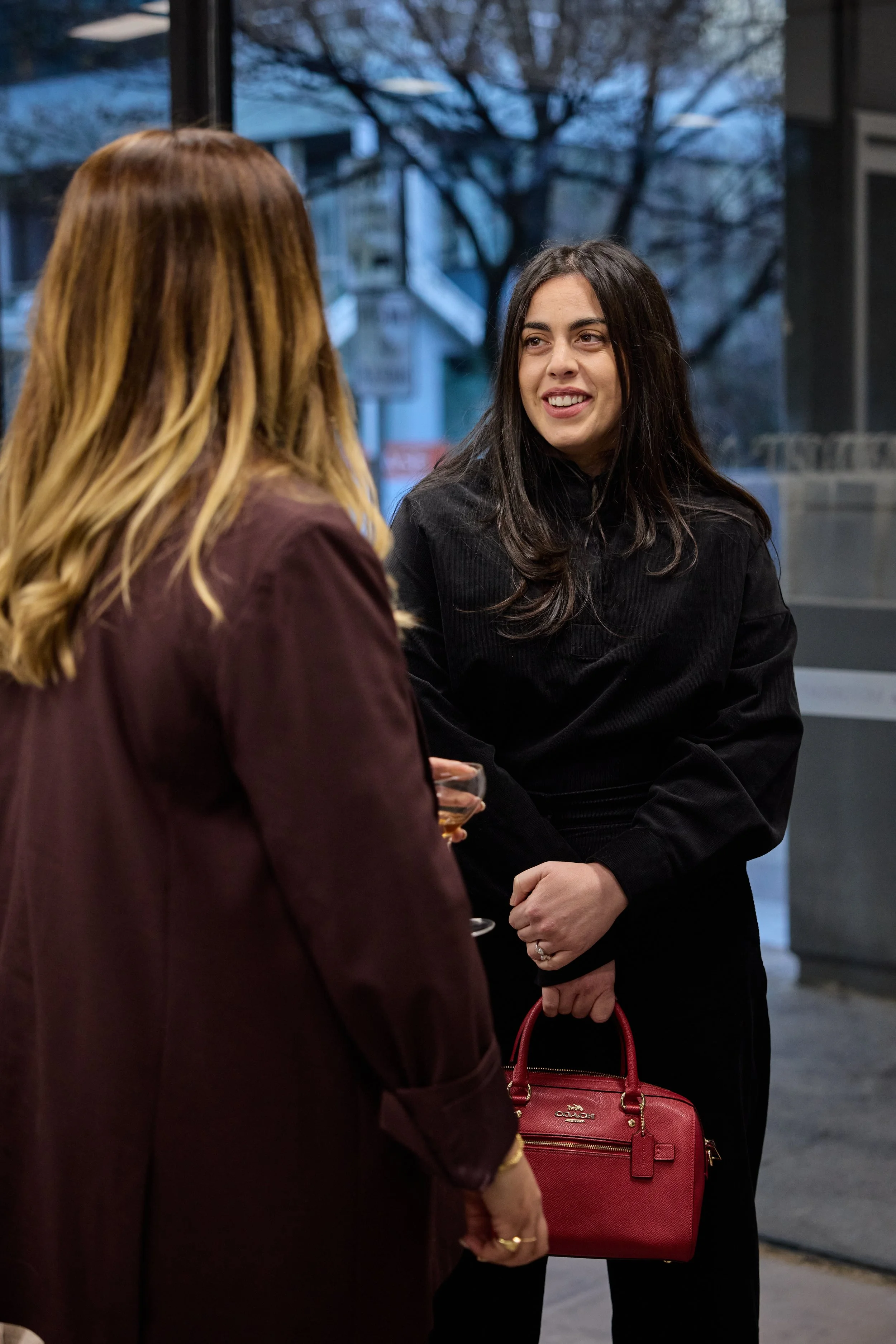 Two women conversing indoors, with one holding a red handbag and a glass of beverage, outside a building with street view and trees in the background.