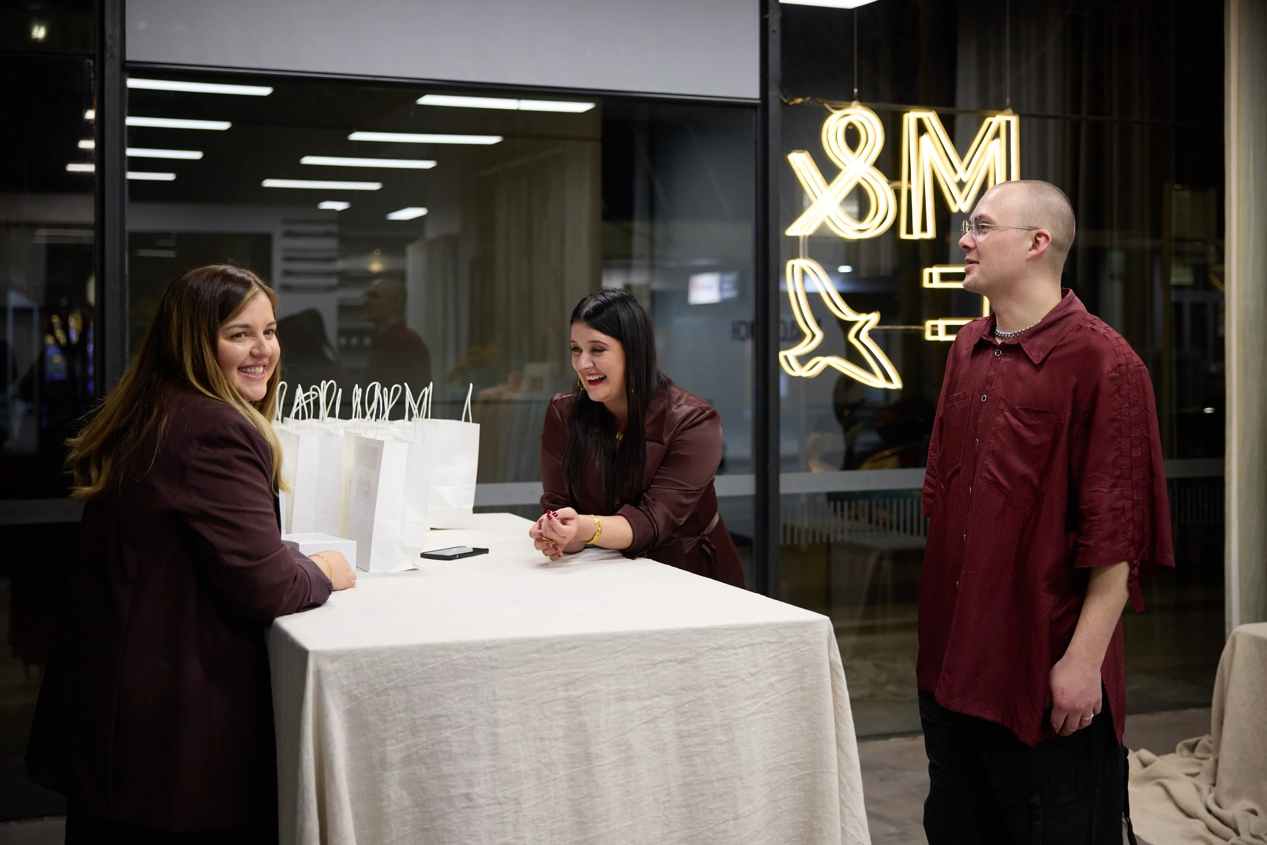 Three people smiling and talking at a registration table in a modern room with glass windows and neon sign.