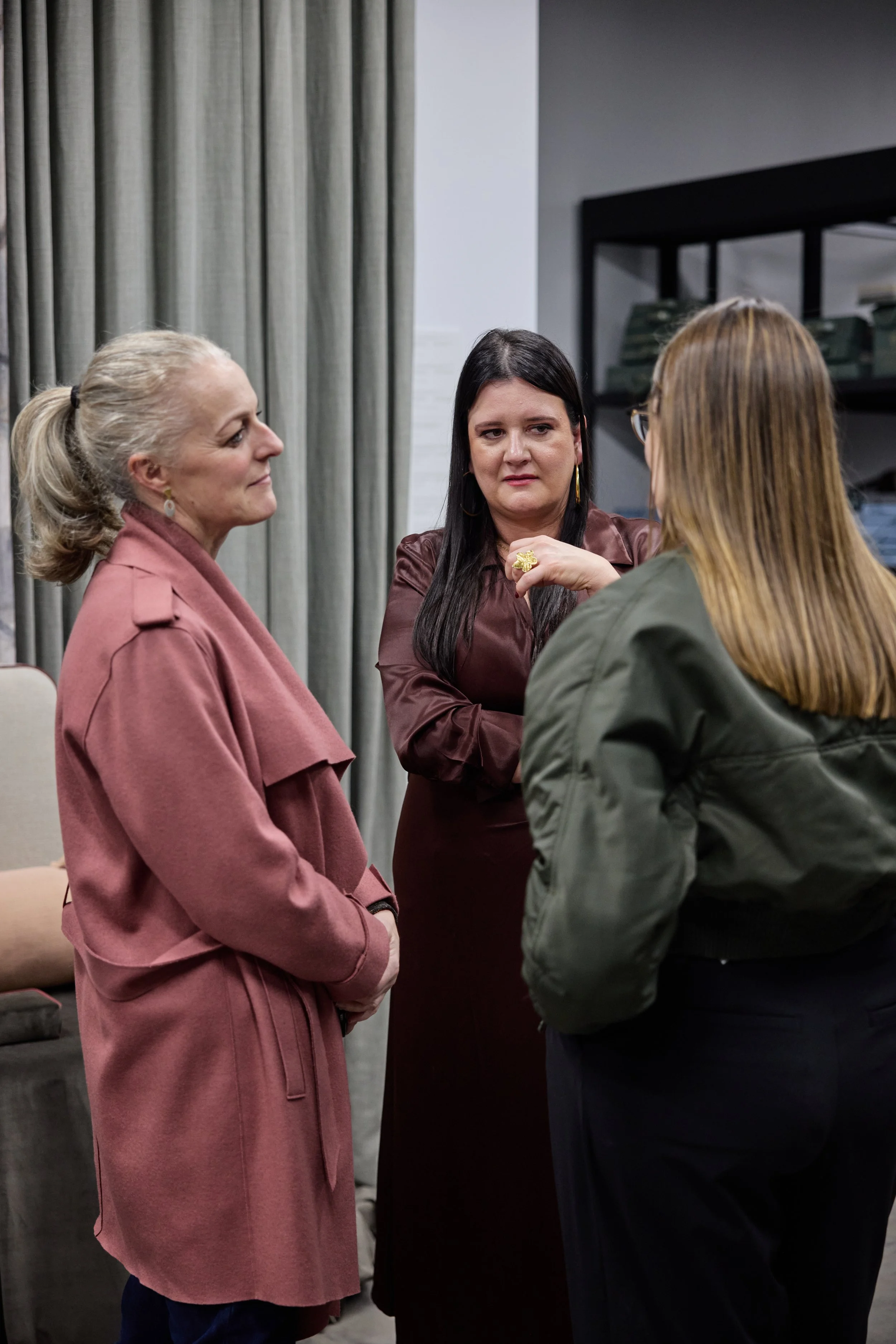 Three women are talking indoors, two with blonde and dark hair, and one with blonde hair and glasses, wearing casual and stylish clothing.