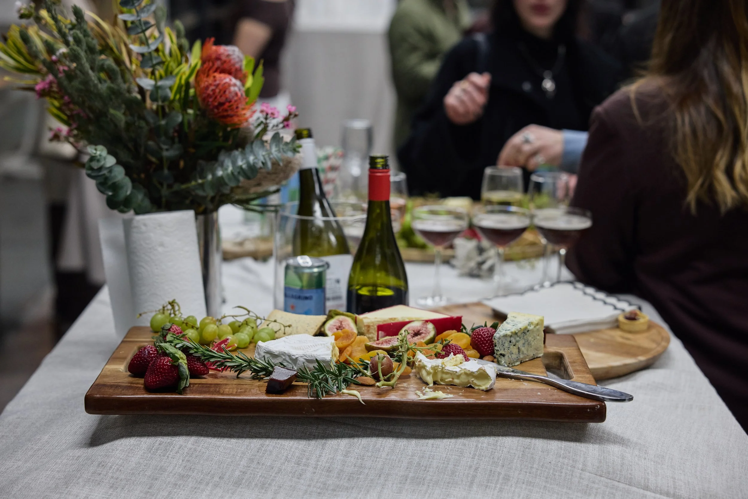 Cheese platter with various cheeses, strawberries, grapes, figs, and herbs on a wooden board, with wine bottles and glasses, and a bouquet of flowers on a table at a social gathering.