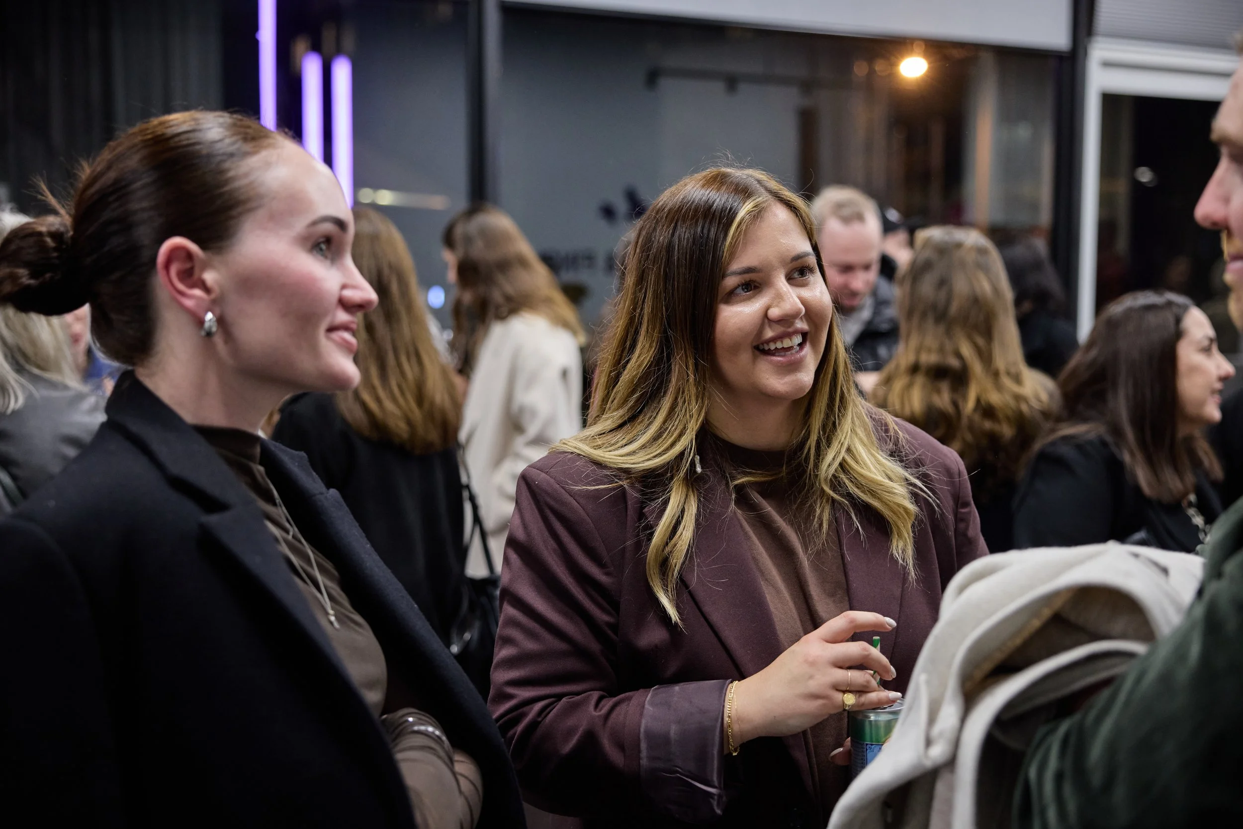 People socializing at an indoor event, with two women in the foreground smiling and talking to each other, surrounded by others in the background.