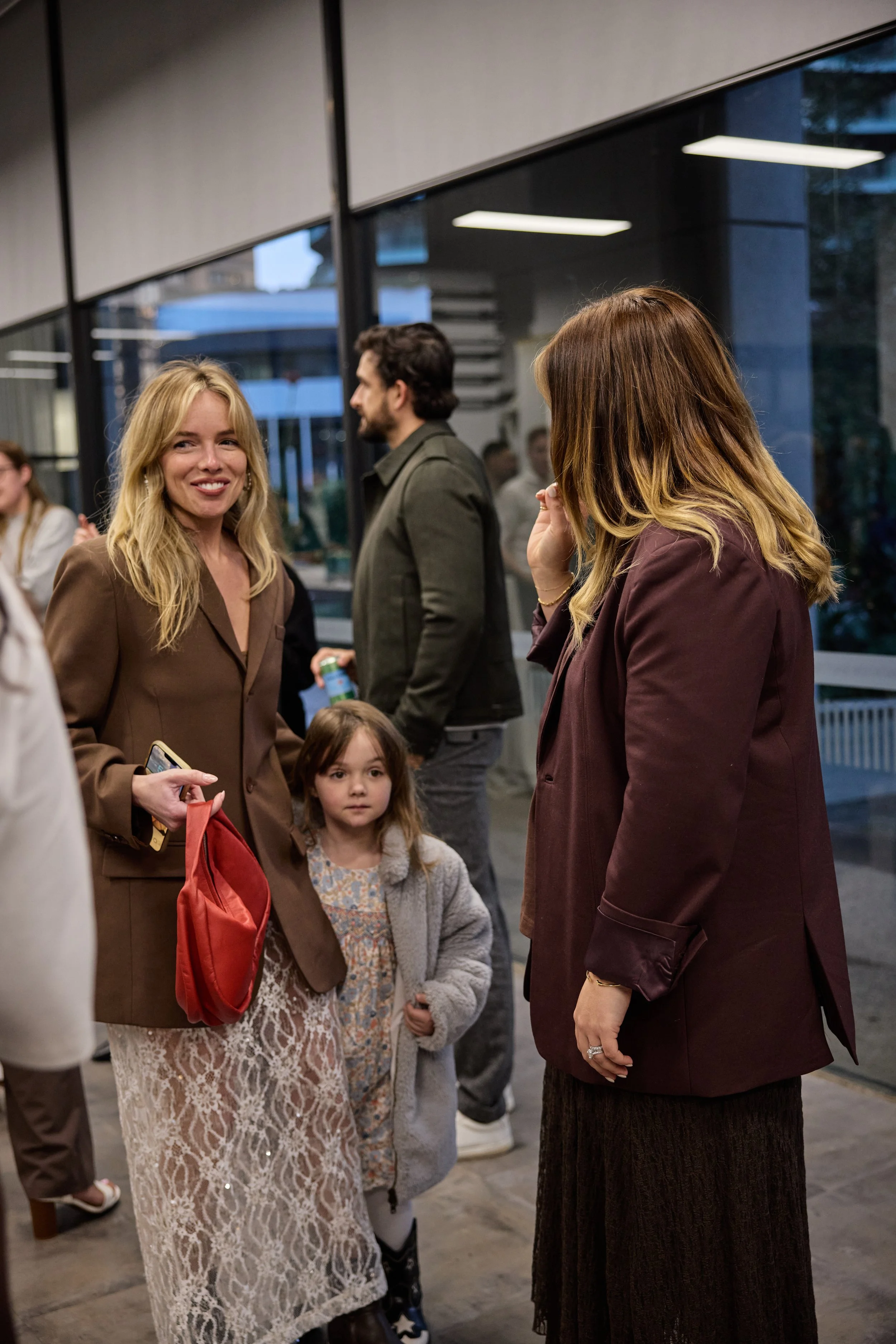 Group of people socializing indoors, including a blonde woman in a brown blazer, a young girl in a patterned dress and gray coat, and a woman with brown hair in a maroon blazer, with others in the background.