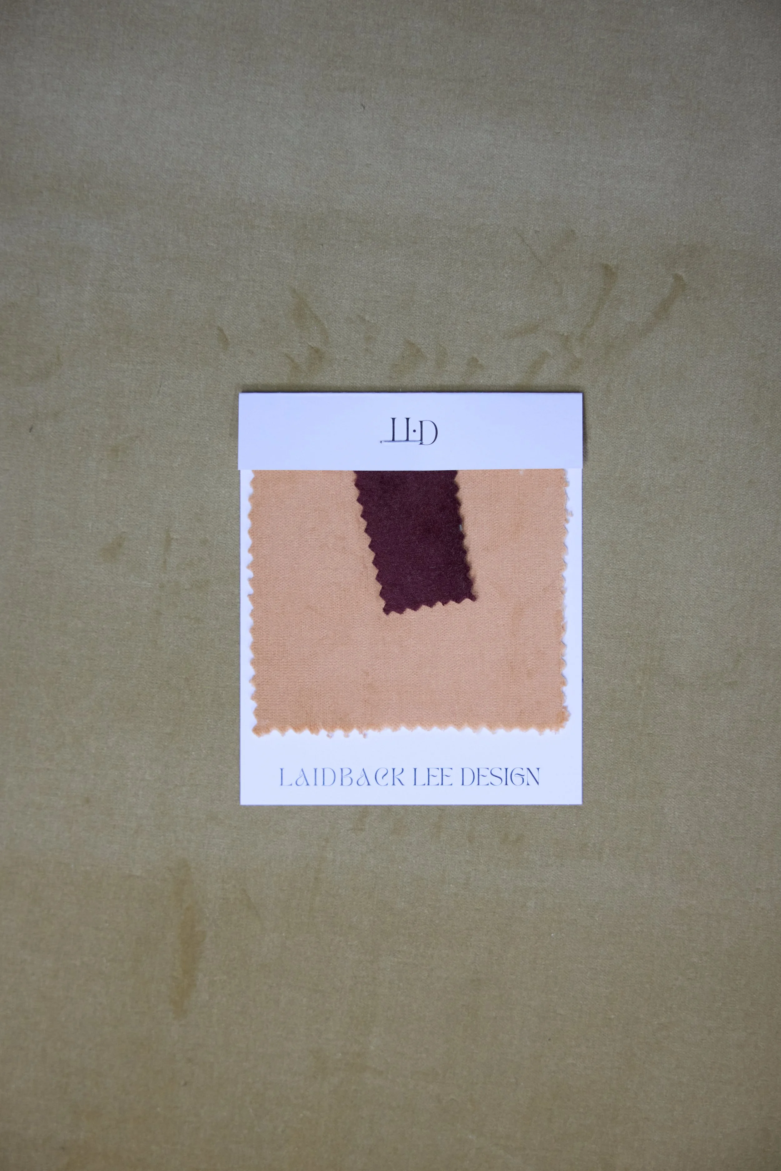 Fabric swatch card with beige and dark brown fabric samples, labeled 'LAIDBACK LEE DESIGN' on white paper background.