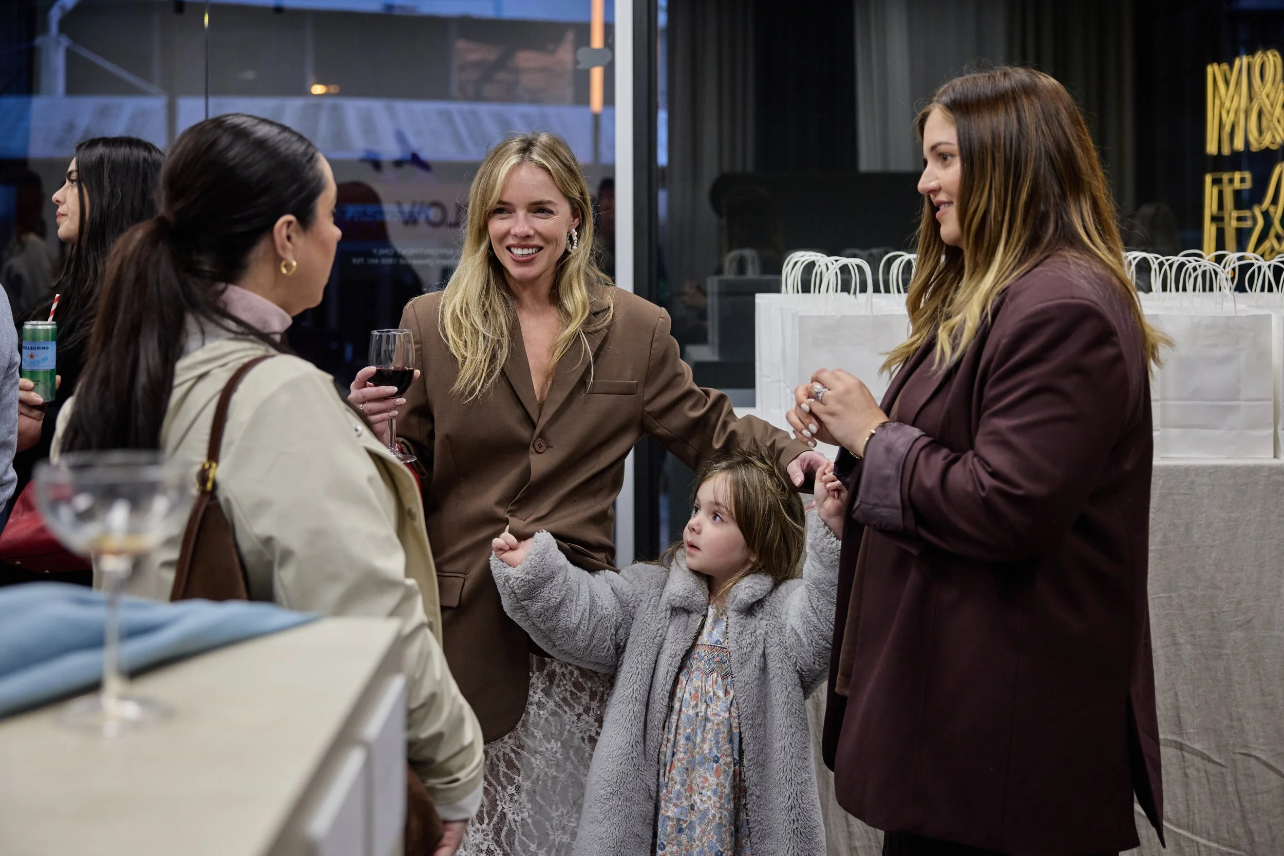 Group of women and a young girl having a conversation at a social gathering indoors, with some holding drinks, surrounded by white shopping bags.
