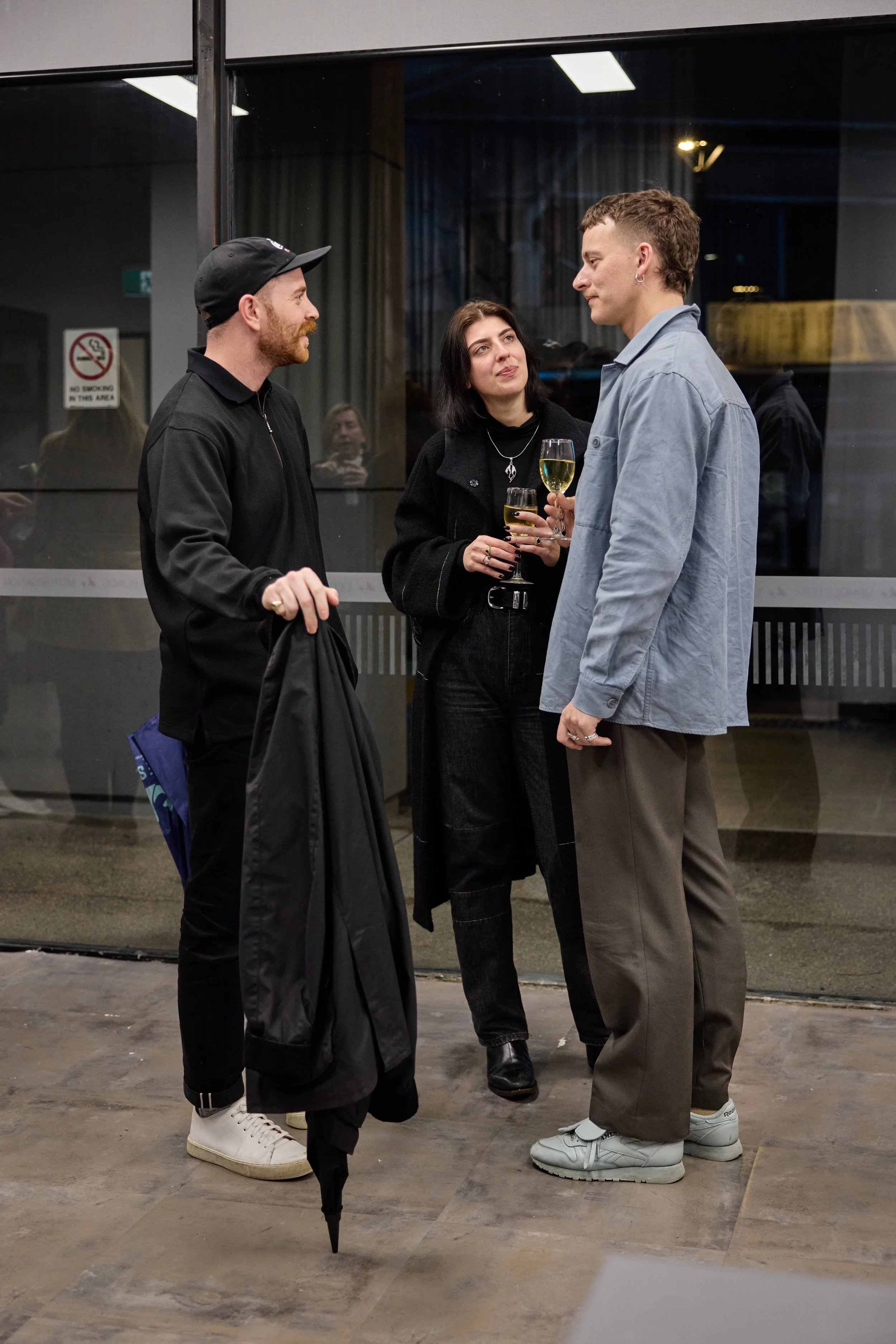 Three people standing outside a building, engaged in conversation. One man holds an umbrella, and two women hold glasses of wine.