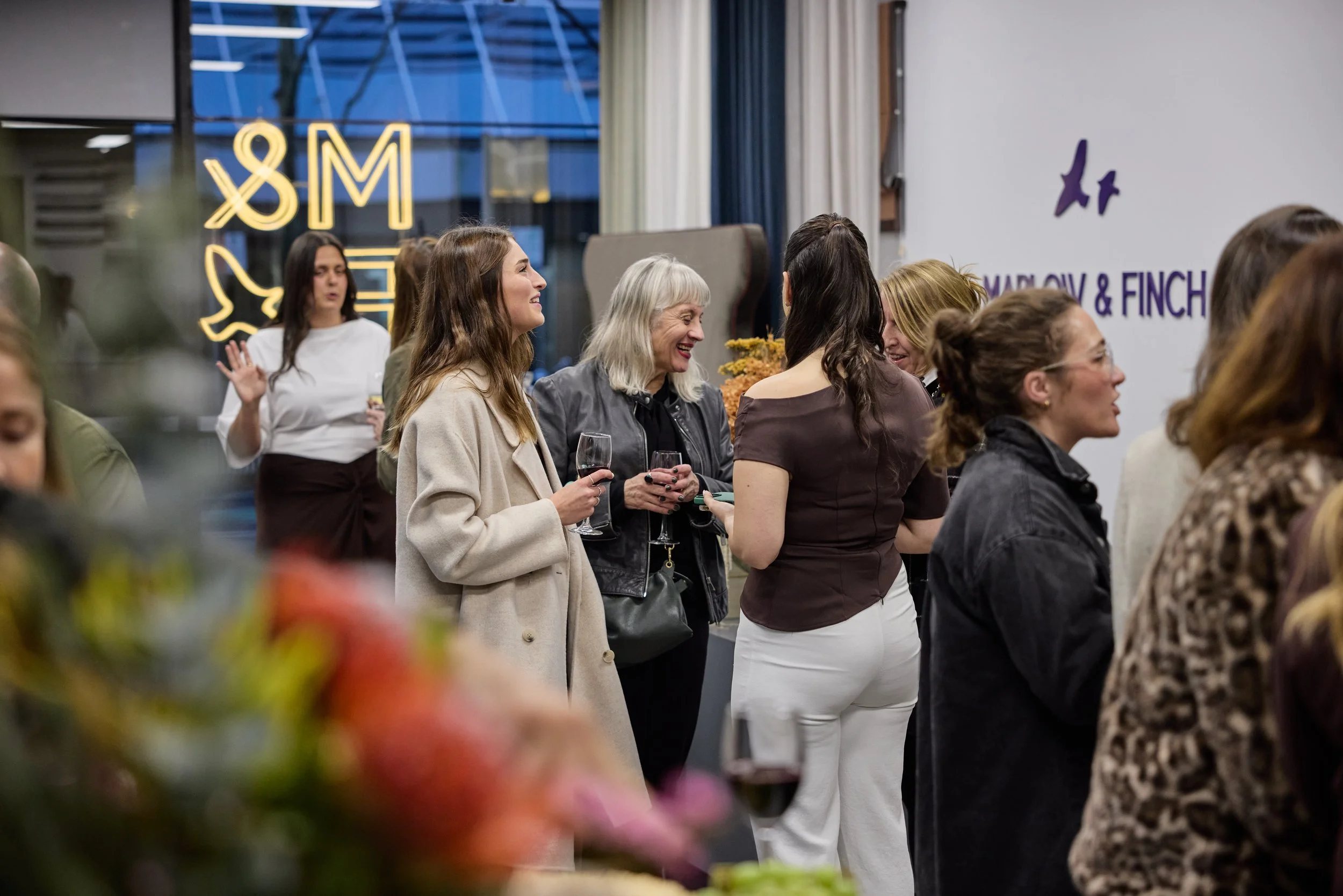 Group of women at a social event, talking and holding glasses of wine, with a sign in the background that reads 'Molly & Finch' and a decorative window with neon letters.