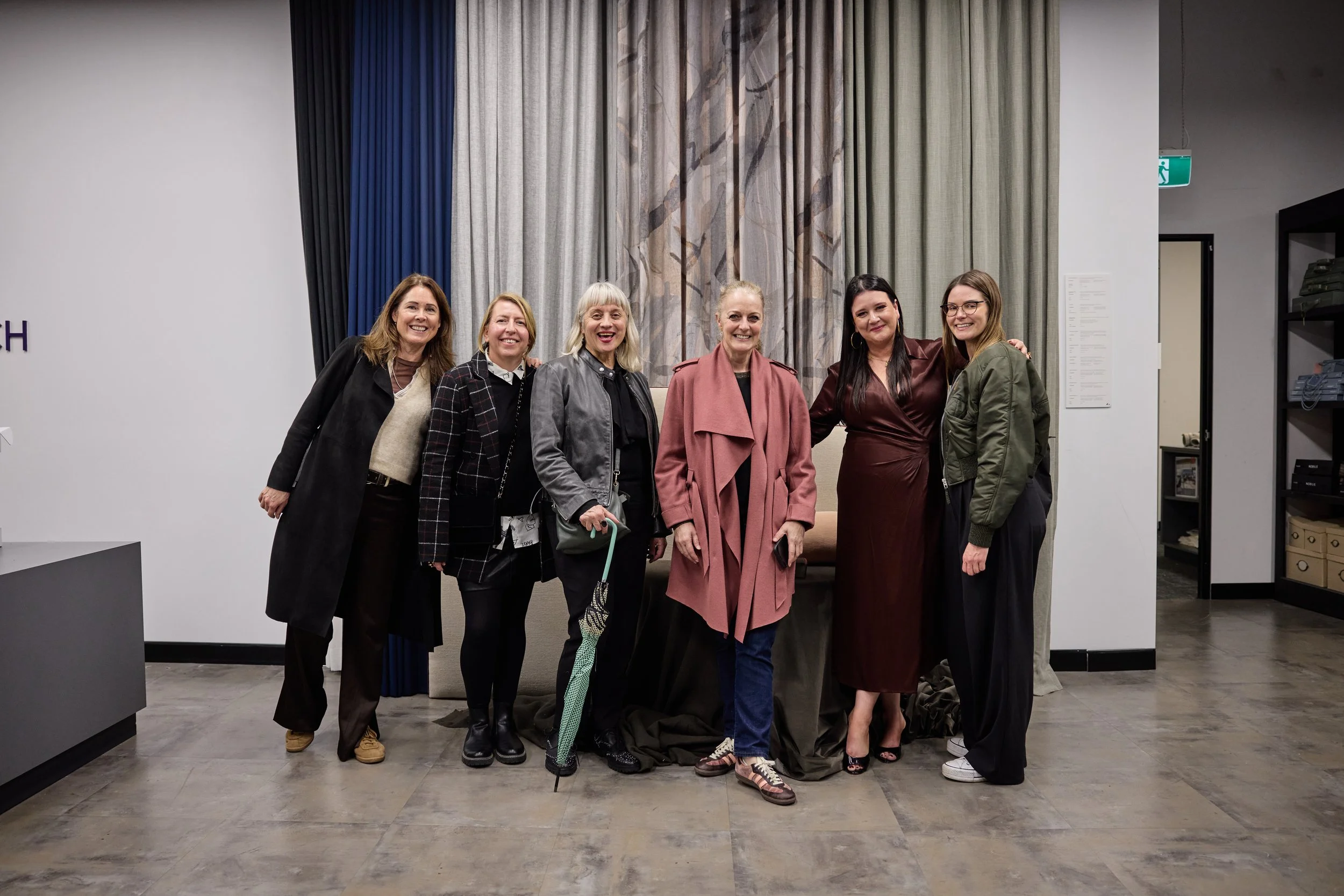 A group of seven women standing together indoors, smiling for a photo, with a background of multicolored curtains and a decorative wall panel.