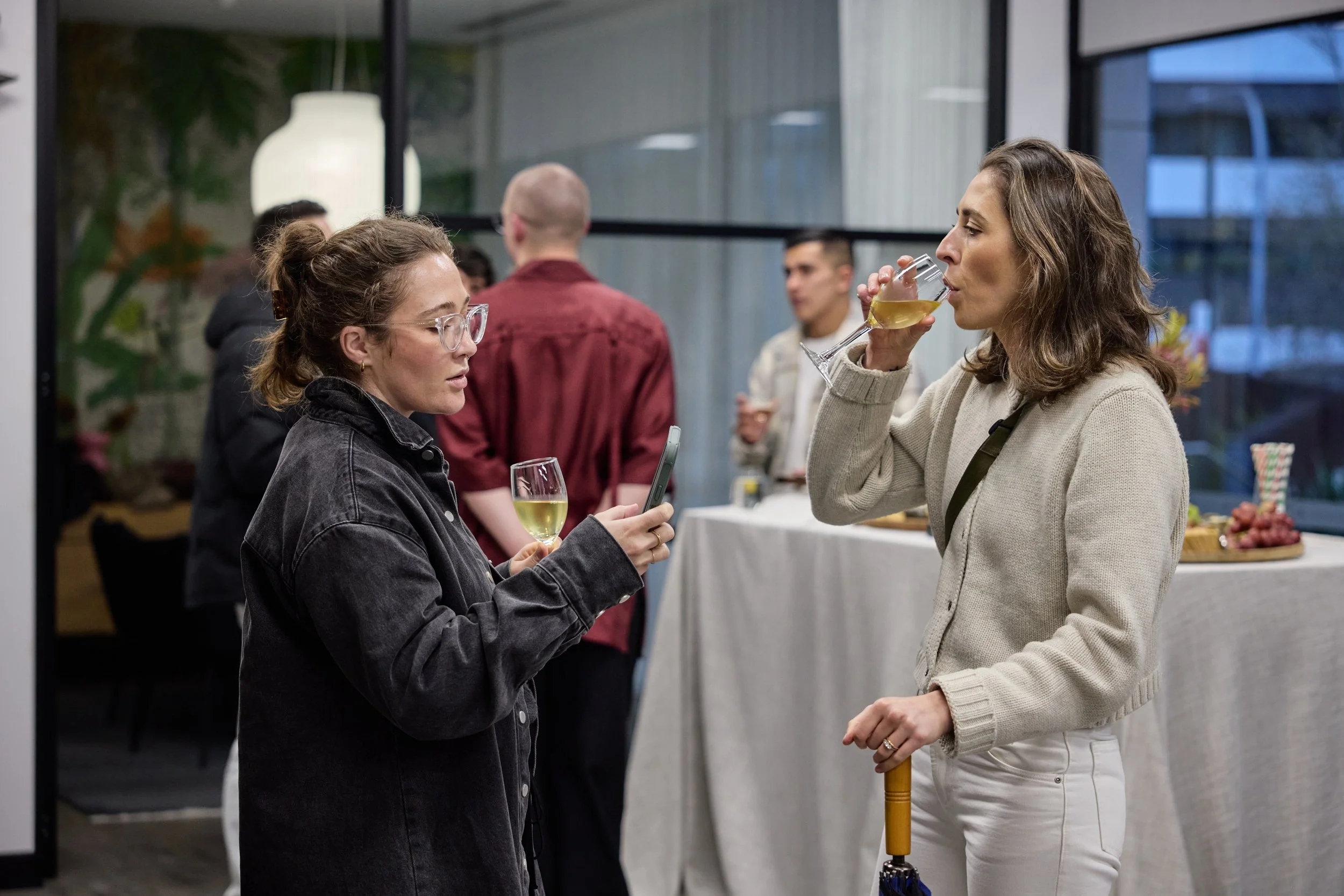 Two women at an indoor social gathering, one holding a wine glass and the other drinking from a glass. Background shows several people and a table with food.