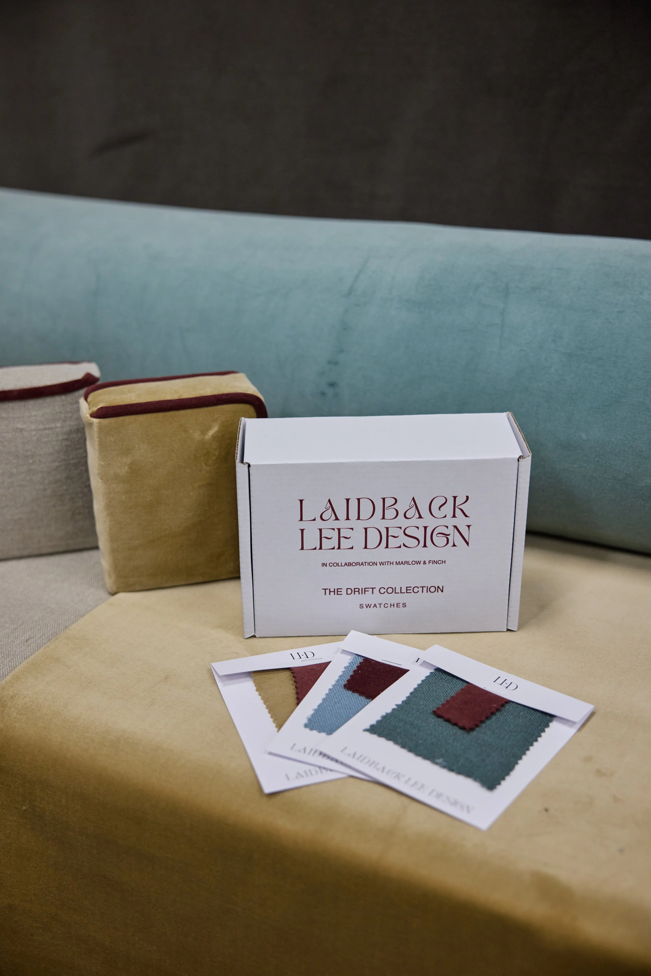 Sample swatches from Laidback Lee Design's Drift Collection, displayed on a beige surface with fabric pillows in background.
