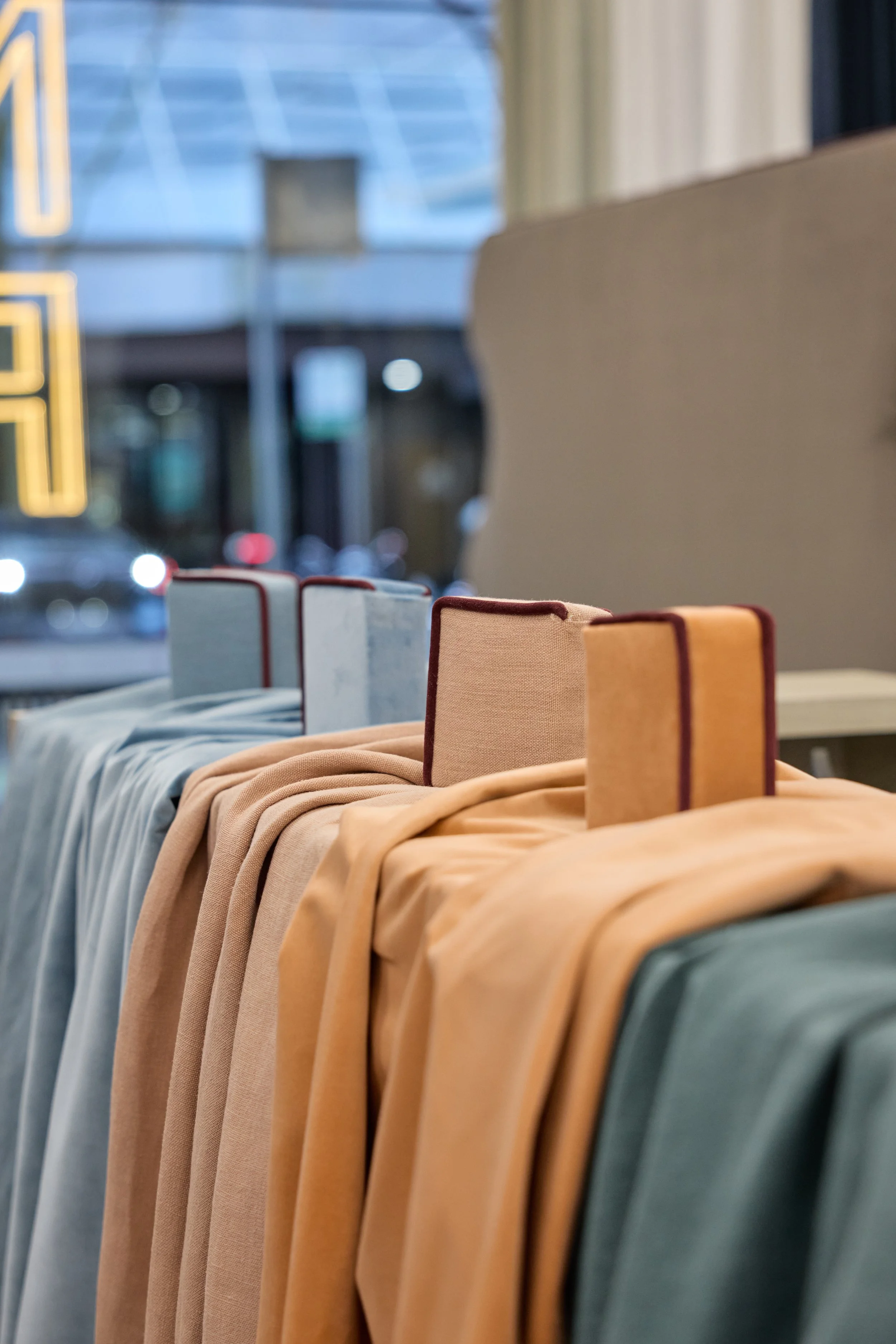Rows of fabric swatches in various colors, with small fabric-covered books standing upright on top, arranged on a table near a window inside a store.