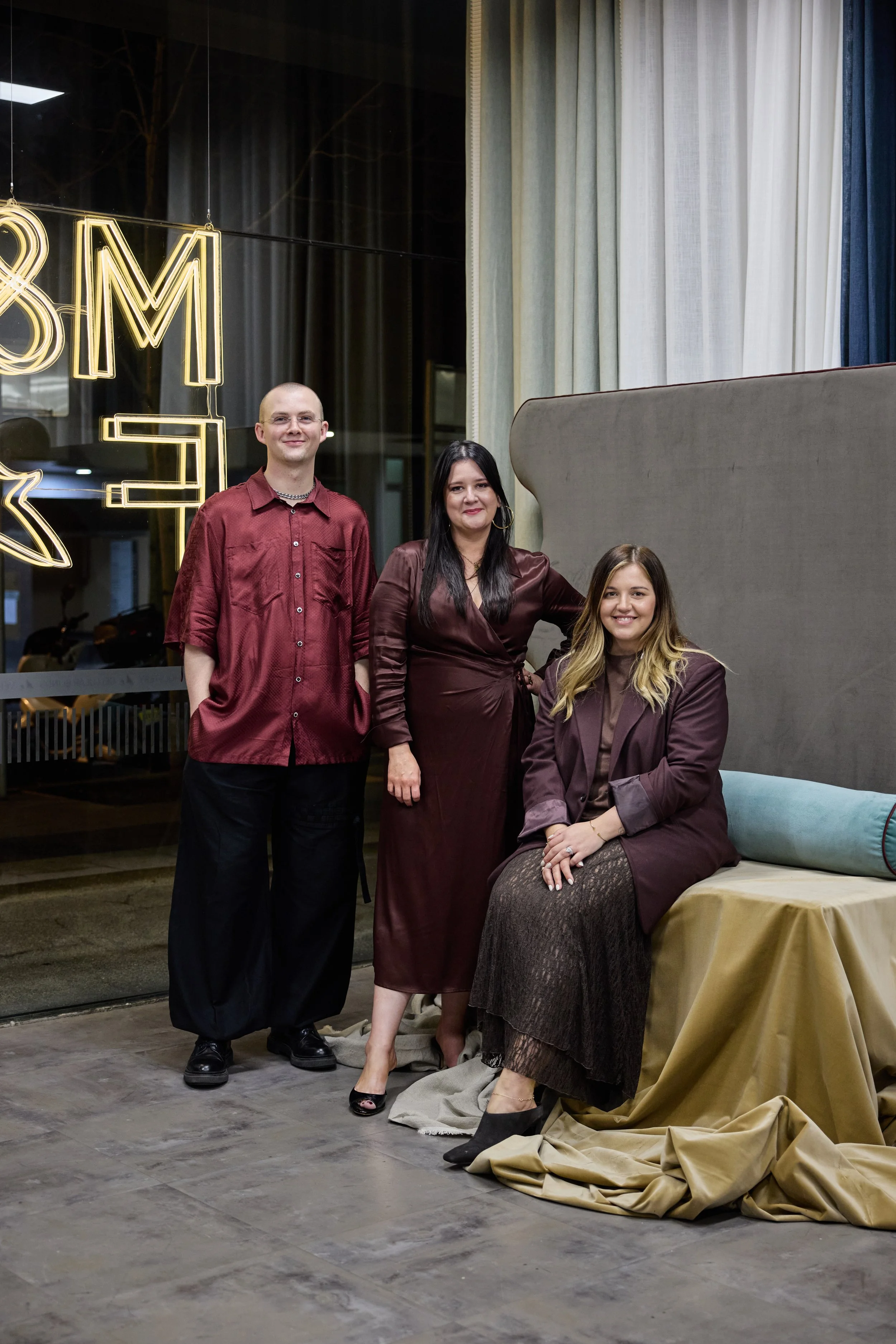 Three people, two women and one man, posing inside a modern venue with large windows, curtains, and a decorative backdrop with neon lights. The women are sitting and the man is standing behind them. They are all dressed in brownish-red clothing.