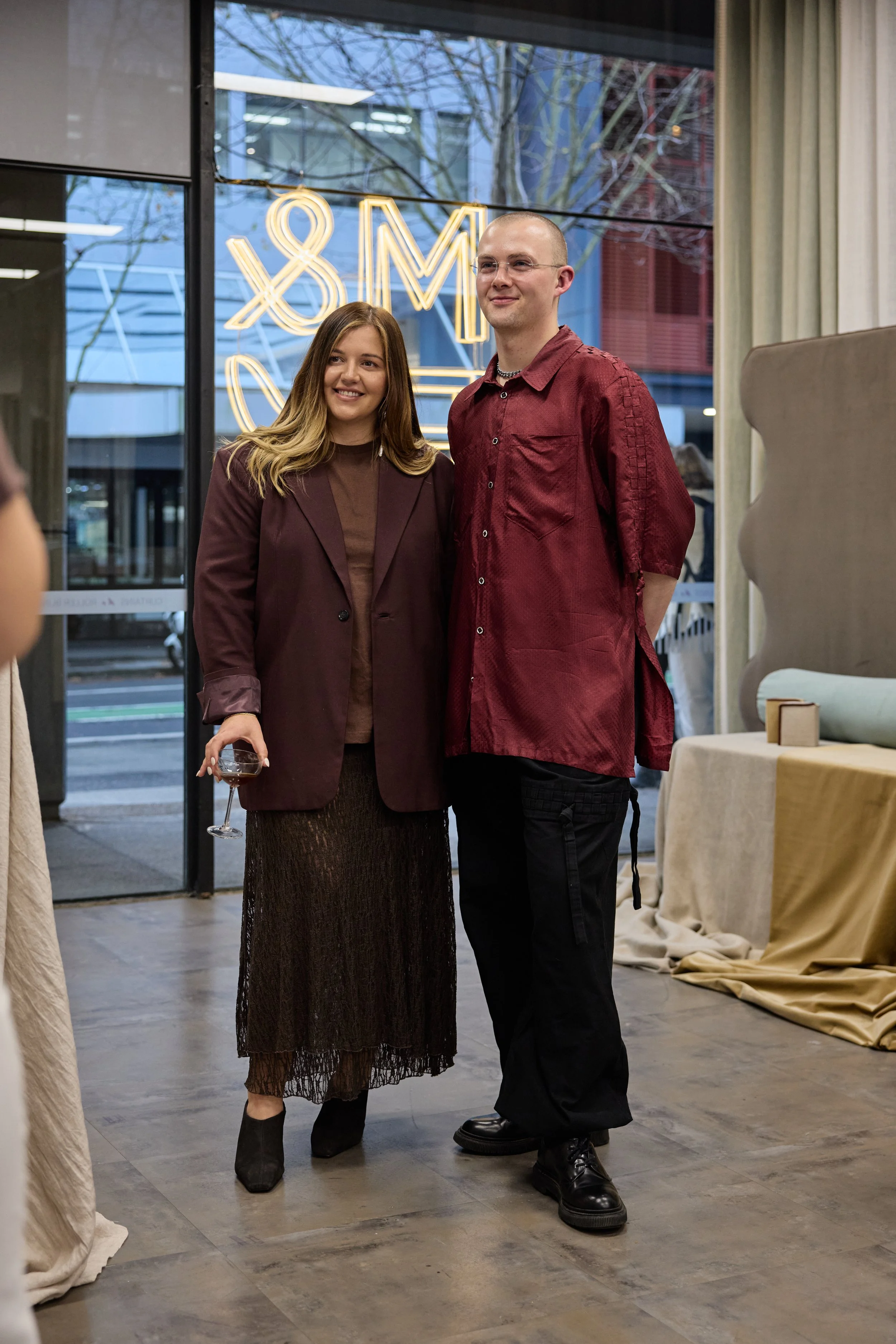 A young woman and a young man stand together inside a store, smiling for a photo. The woman holds a glass of red wine and wears a brown skirt, a maroon blazer, and black shoes. The man wears a red shirt, black pants, and black shoes. Behind them, a l