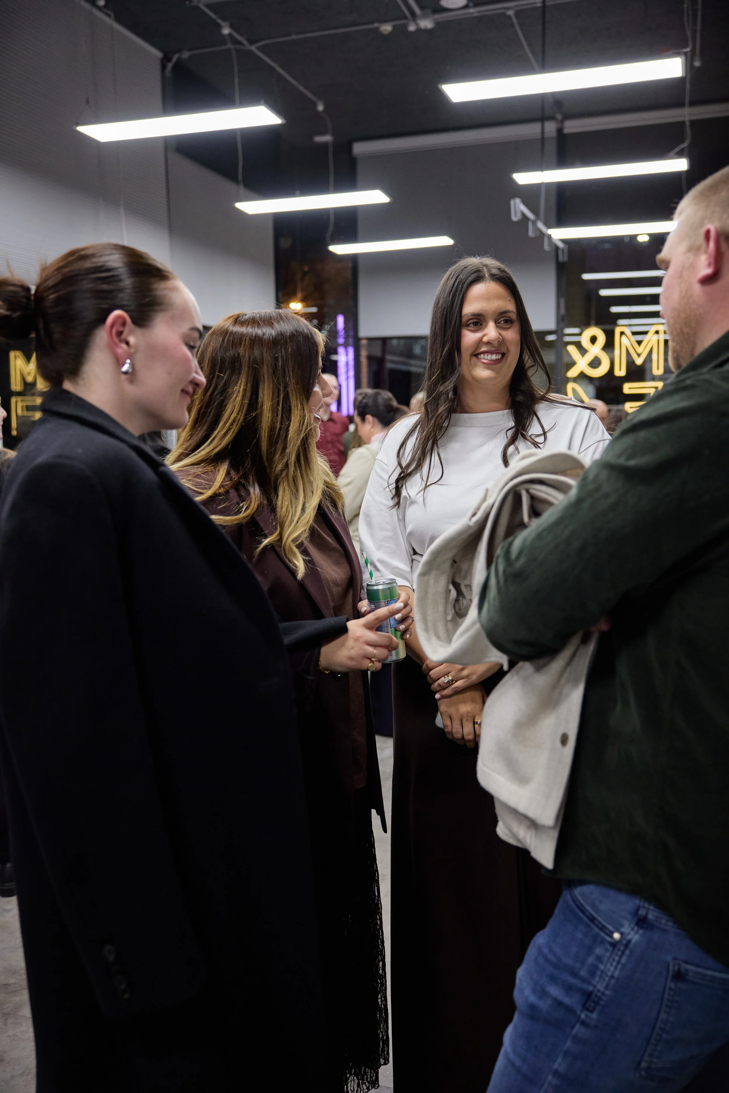 Group of young adults socializing at a modern indoor event, with women smiling and talking, holding drinks, and a man with crossed arms.