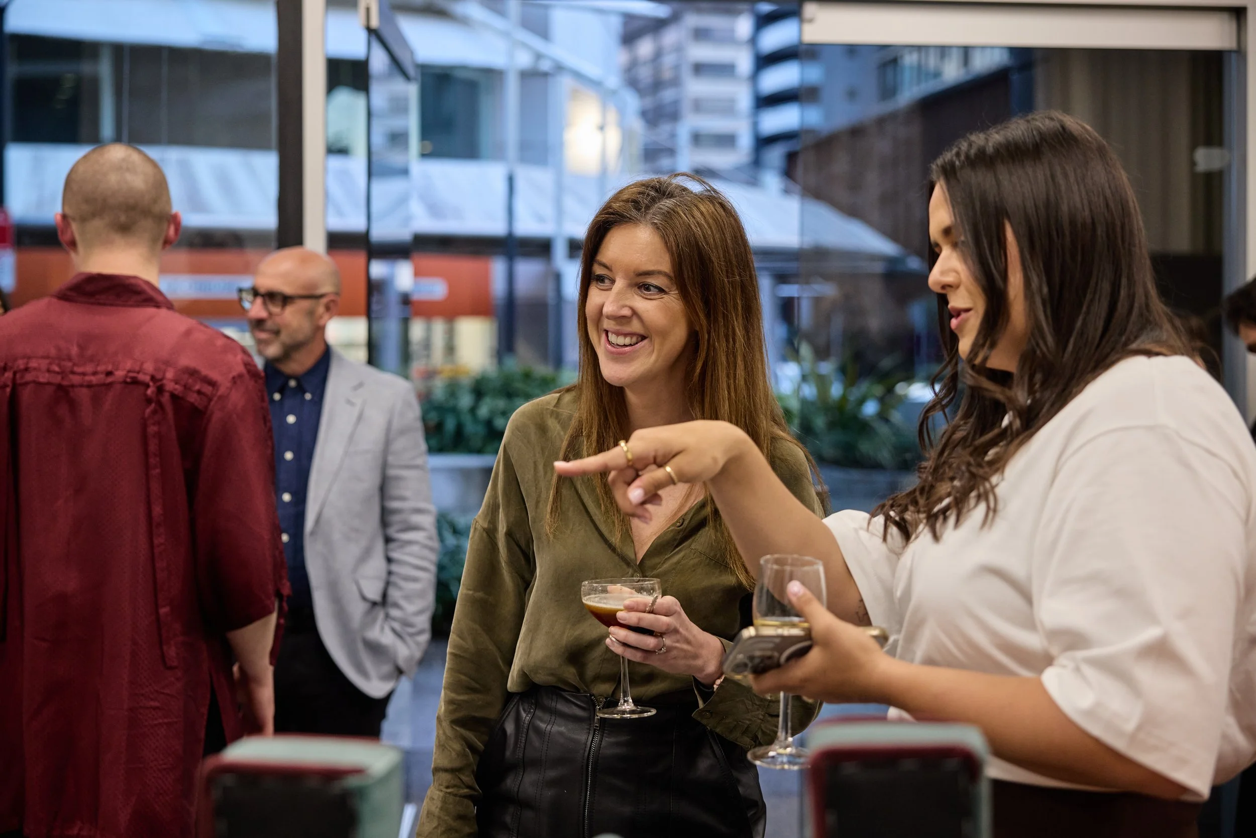 Group of people at a social gathering, talking and smiling, with drinks in hand, outside of a building during daytime.