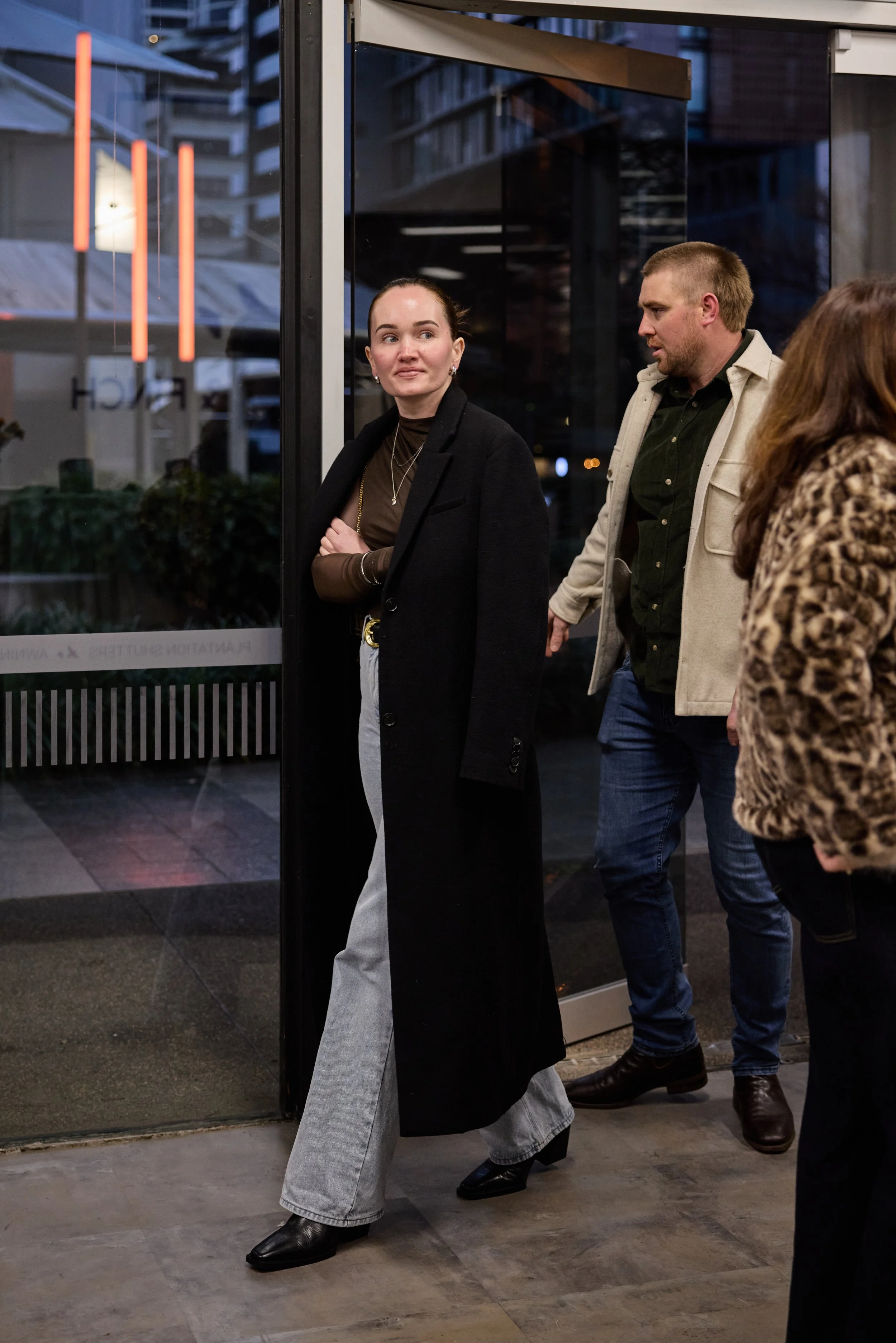 A woman in a long black coat and light-colored jeans walking out of a building while two men and a woman follow behind her in a city setting at dusk.