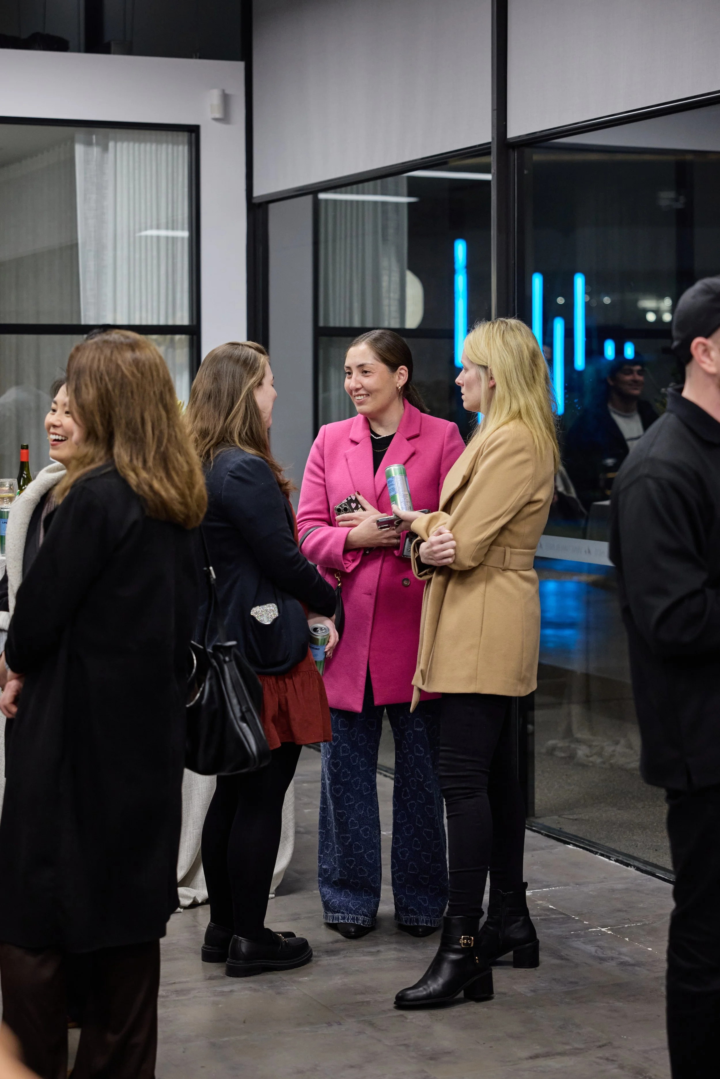 Group of women chatting at an indoor social event, some holding drinks, wearing stylish coats, with large glass windows in the background and blue neon lights outside.