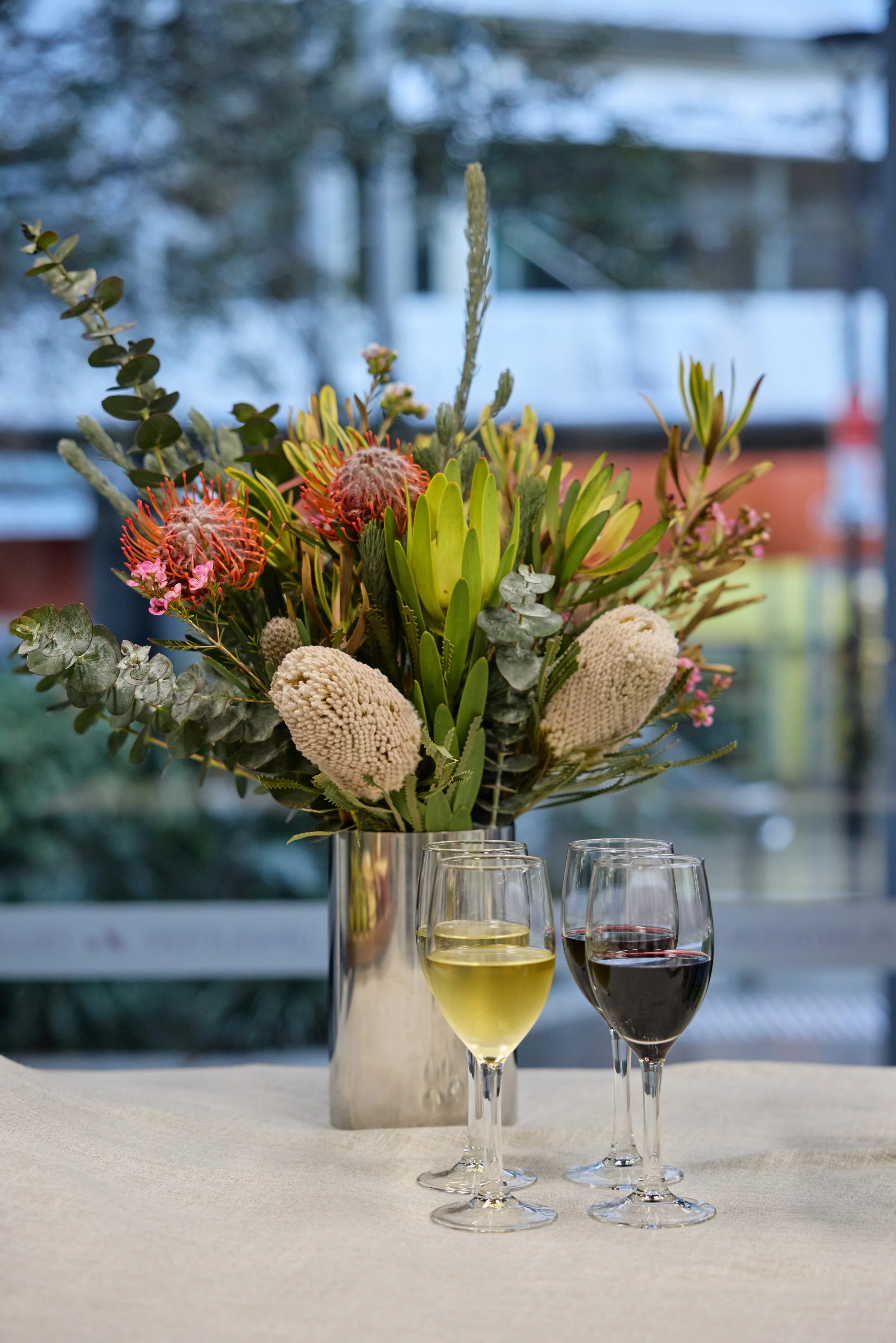 A large floral arrangement with various flowers and greenery in a silver vase, placed behind three glasses of wine on a table with a light-colored tablecloth. Two glasses contain white wine, and one contains red wine.