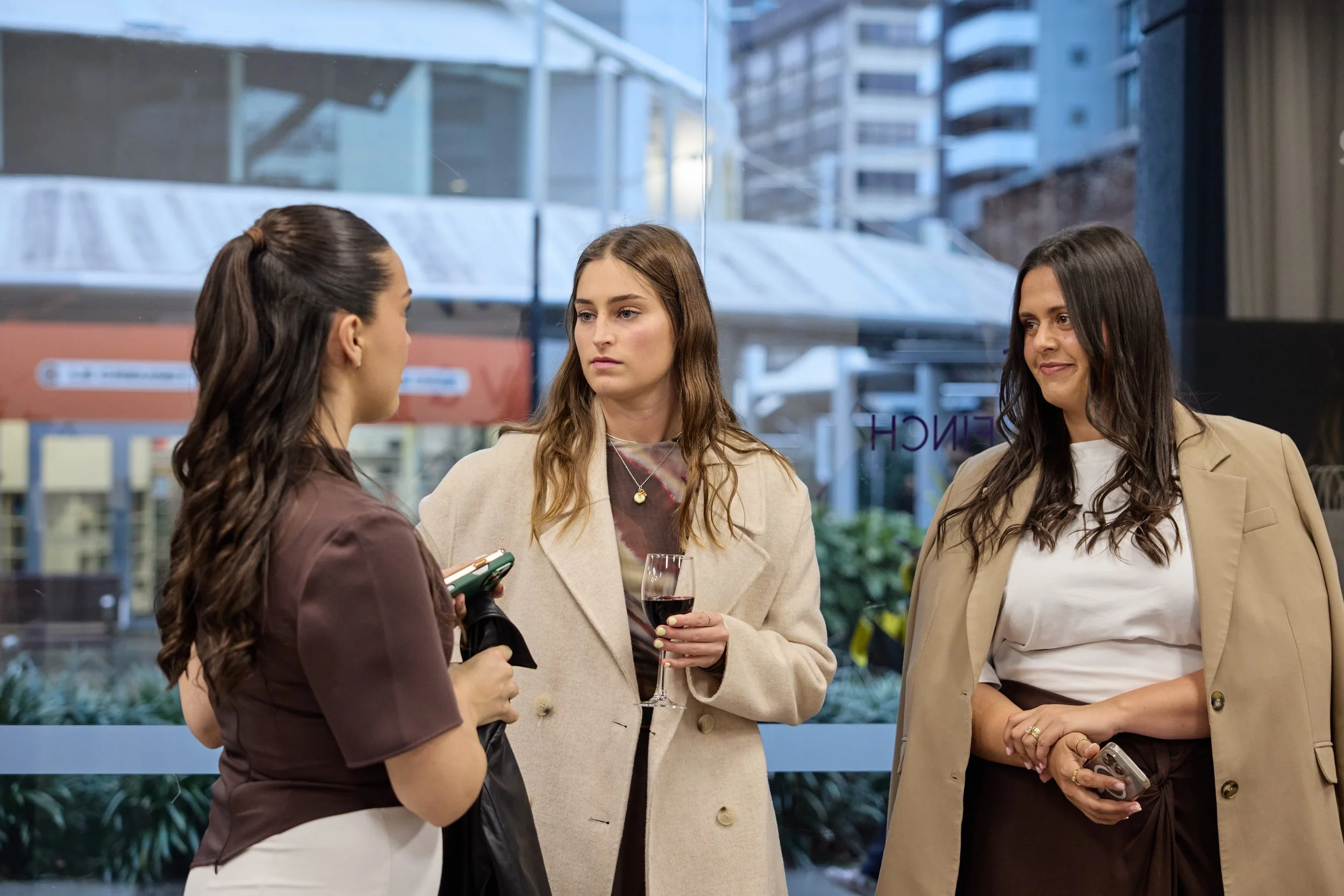 Three women standing and talking inside a building near a large window with city buildings outside. The woman on the left has long dark hair tied in a ponytail and is wearing a brown top, holding a smartphone. The woman in the center has light brown
