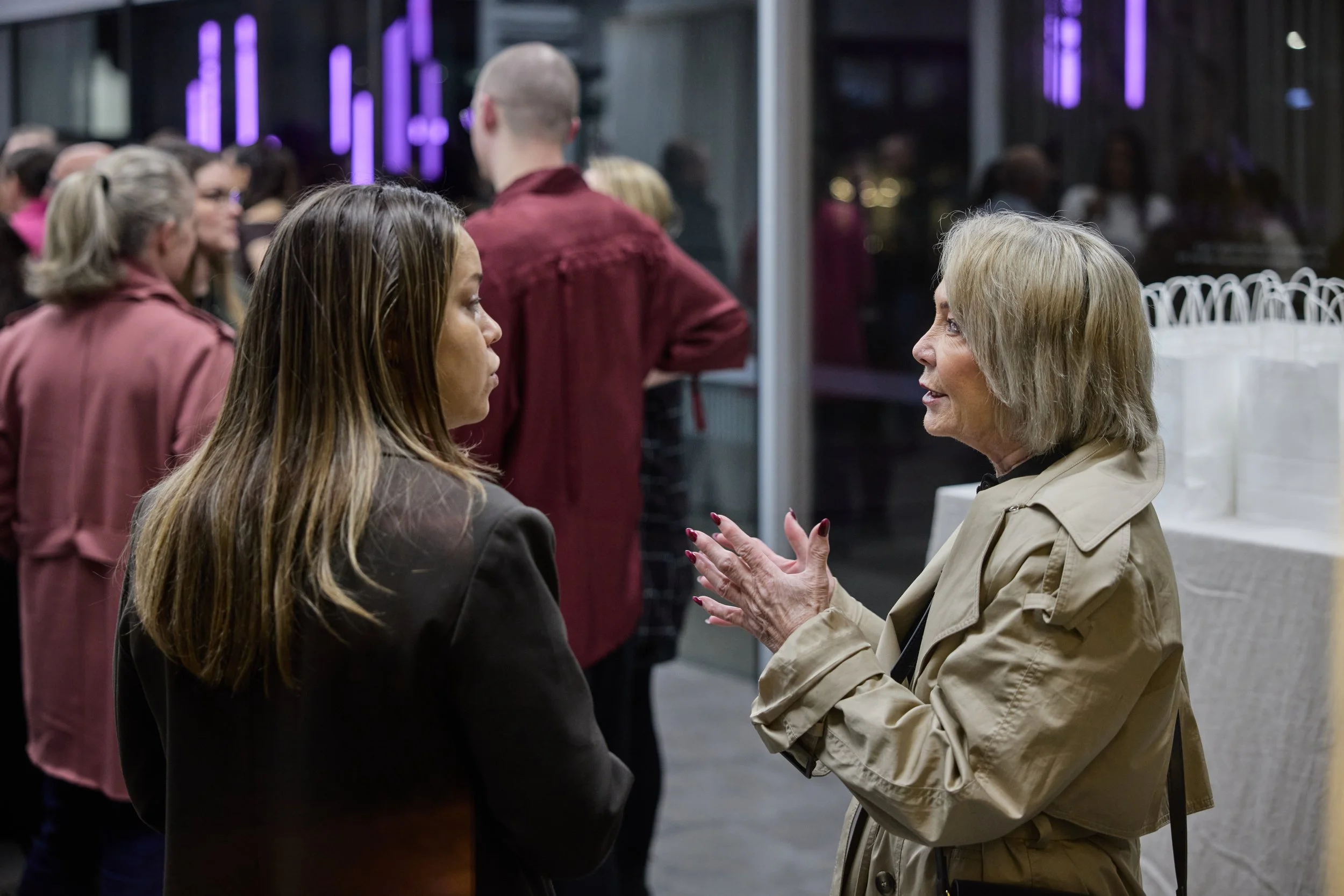 Two women are engaged in conversation at a social event, with a group of people in the background.
