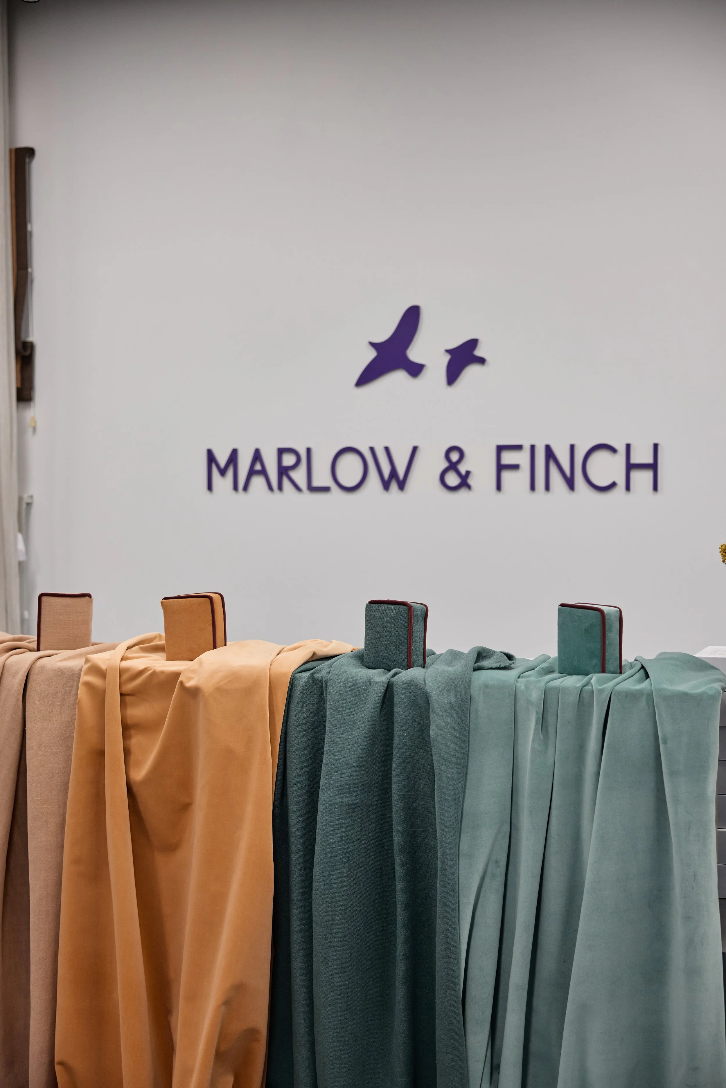 Display of fabric swatches in various shades of beige, orange, teal, and green, each draped over a stand in front of a white wall with the Marlow & Finch logo and birds