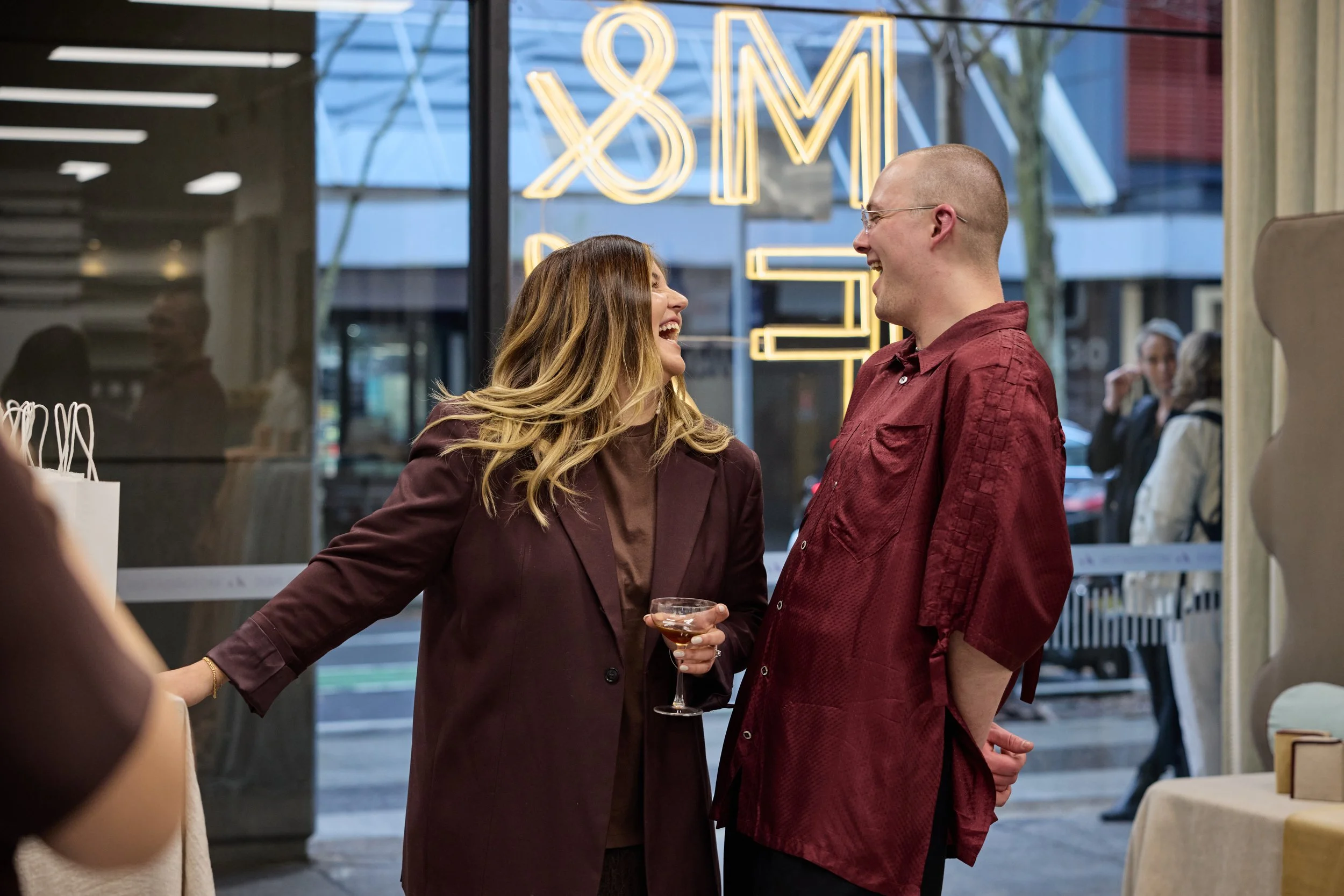 Two people, a woman with long wavy hair and a man with short hair and glasses, are laughing and talking inside a store or restaurant. The woman is holding a glass of wine, and they're standing near a window with a neon sign behind them that is partia