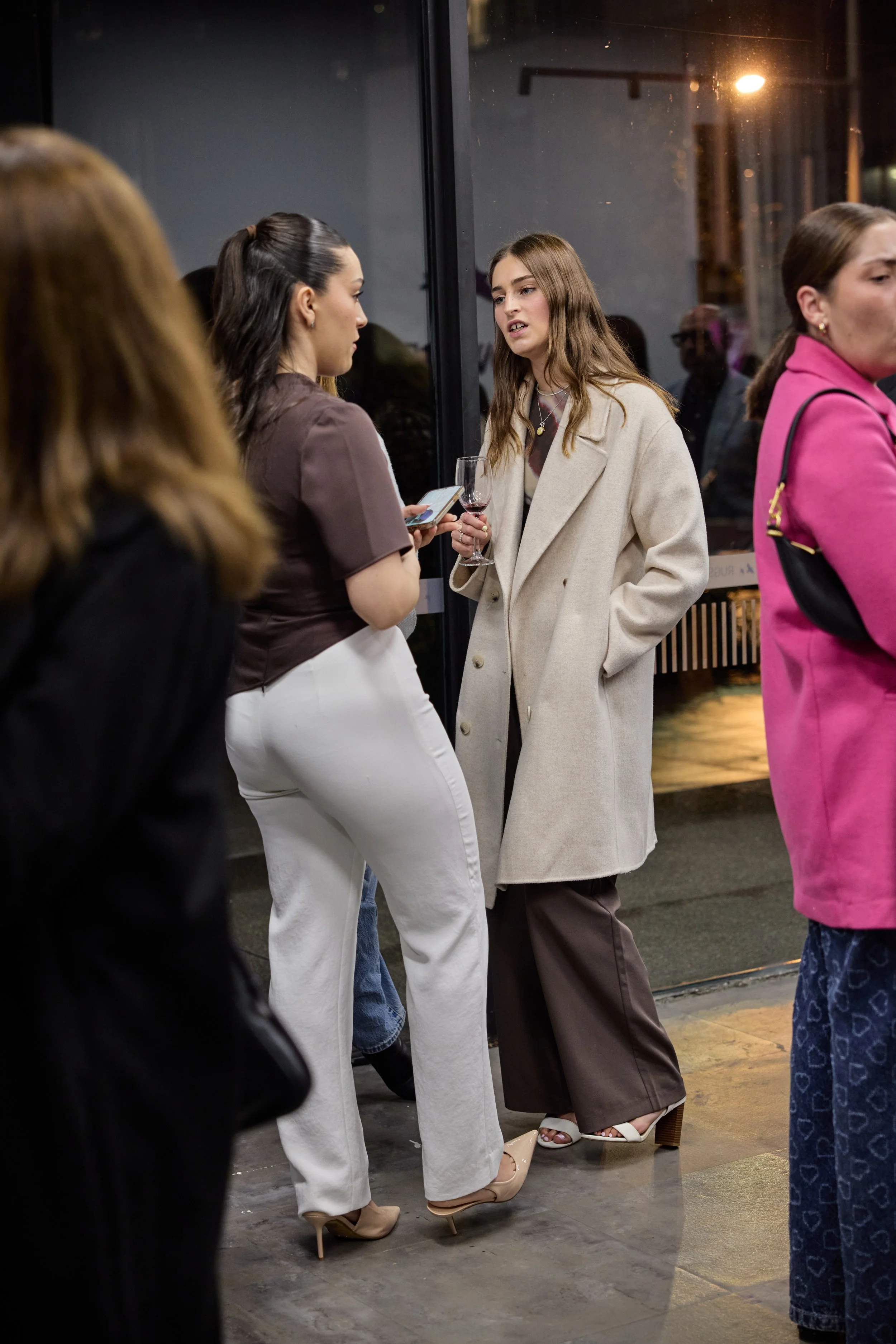 Two women are engaged in a conversation outside a building during the night, one holding a glass of wine, with other people nearby.