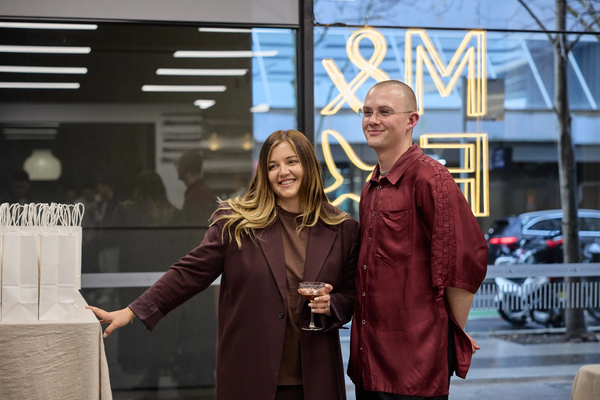 Two people, a woman with long light brown hair and a man with a shaved head, standing inside a modern building with large windows and neon signs in the background. The woman is smiling and holding a cocktail glass, while the man is smiling with his h