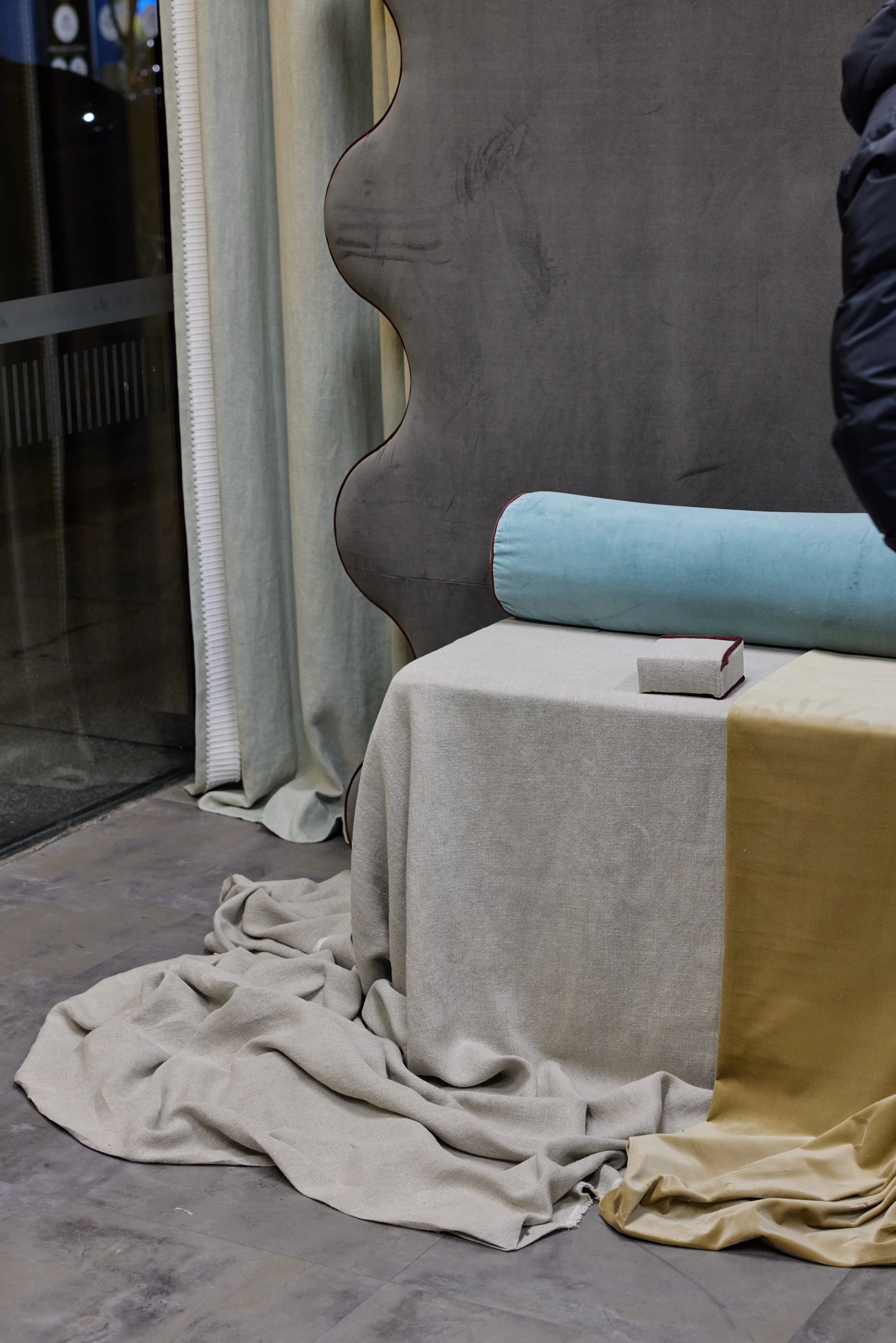 Interior scene with a table draped in beige and gold fabrics, a blue cylindrical pillow, a small gray box, and a decorative wavy wooden partition near a glass door with curtains.