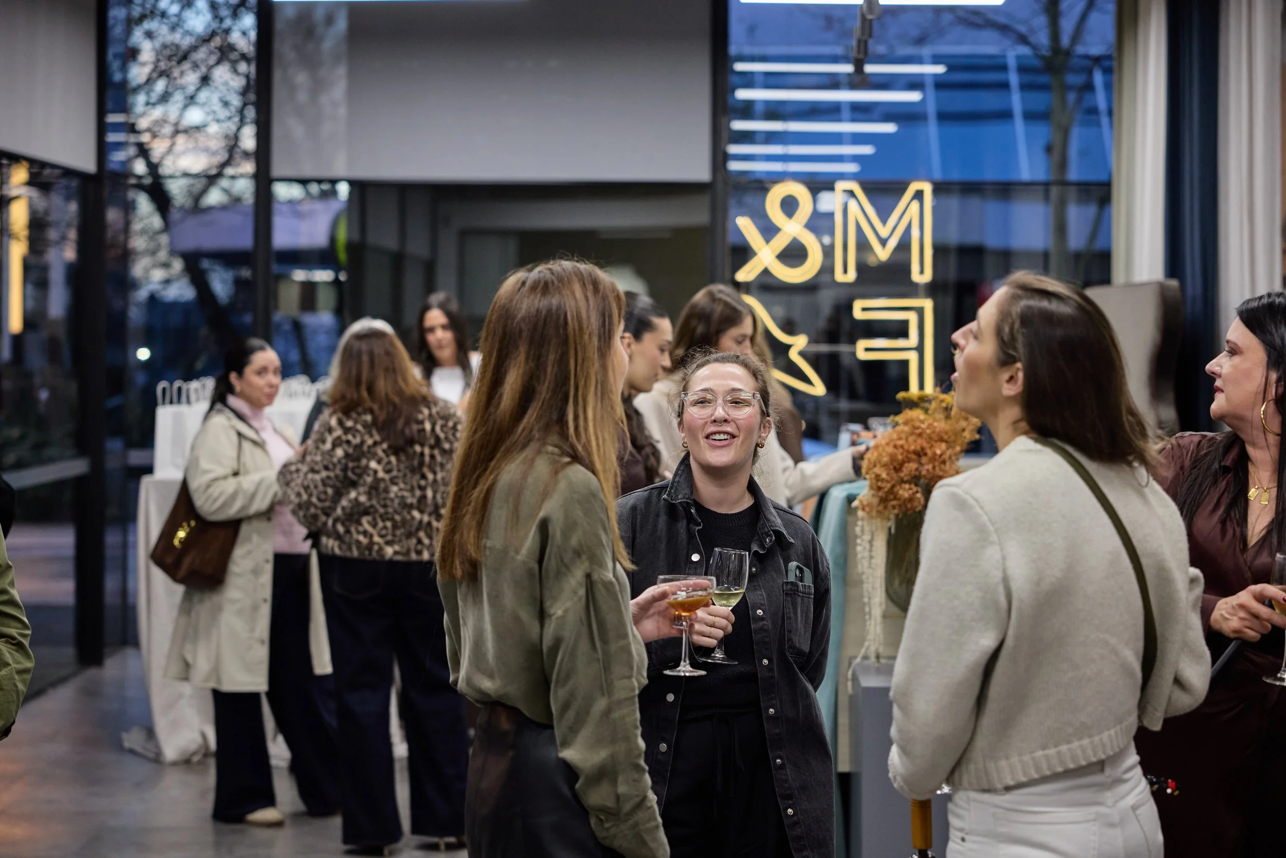 Group of women socializing at a lively indoor gathering or party, some holding glasses of wine or champagne, in a modern venue with glass windows and neon signage.