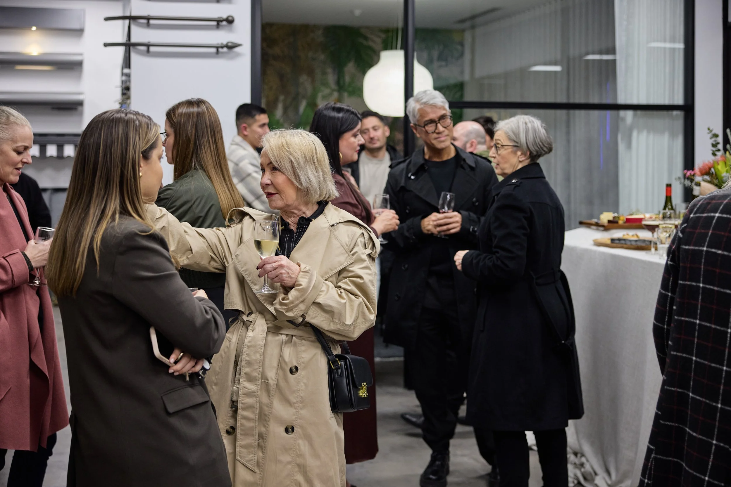 People mingling at a social gathering or party, some holding glasses of wine, in an indoor setting with modern decor and a large window in the background.