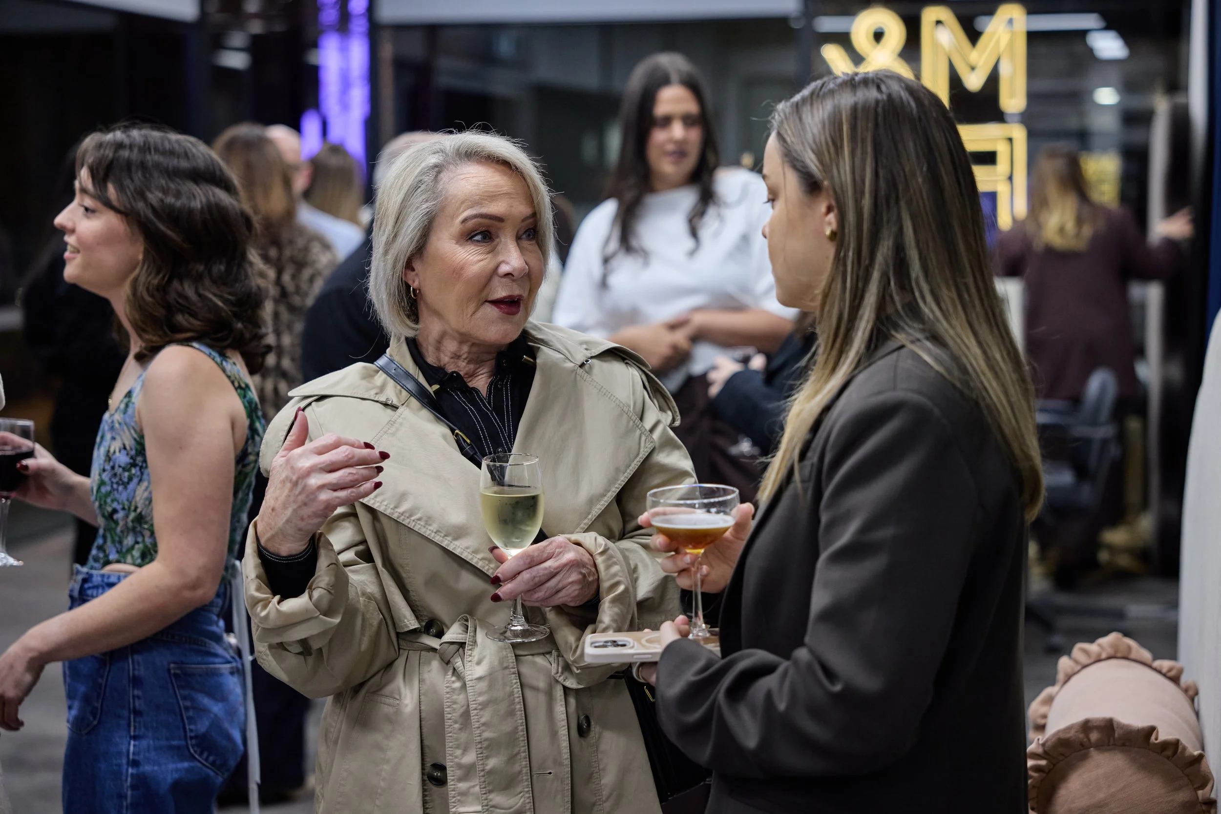 Two women engaging in conversation at a social gathering, each holding a glass of wine, with other people in the background.