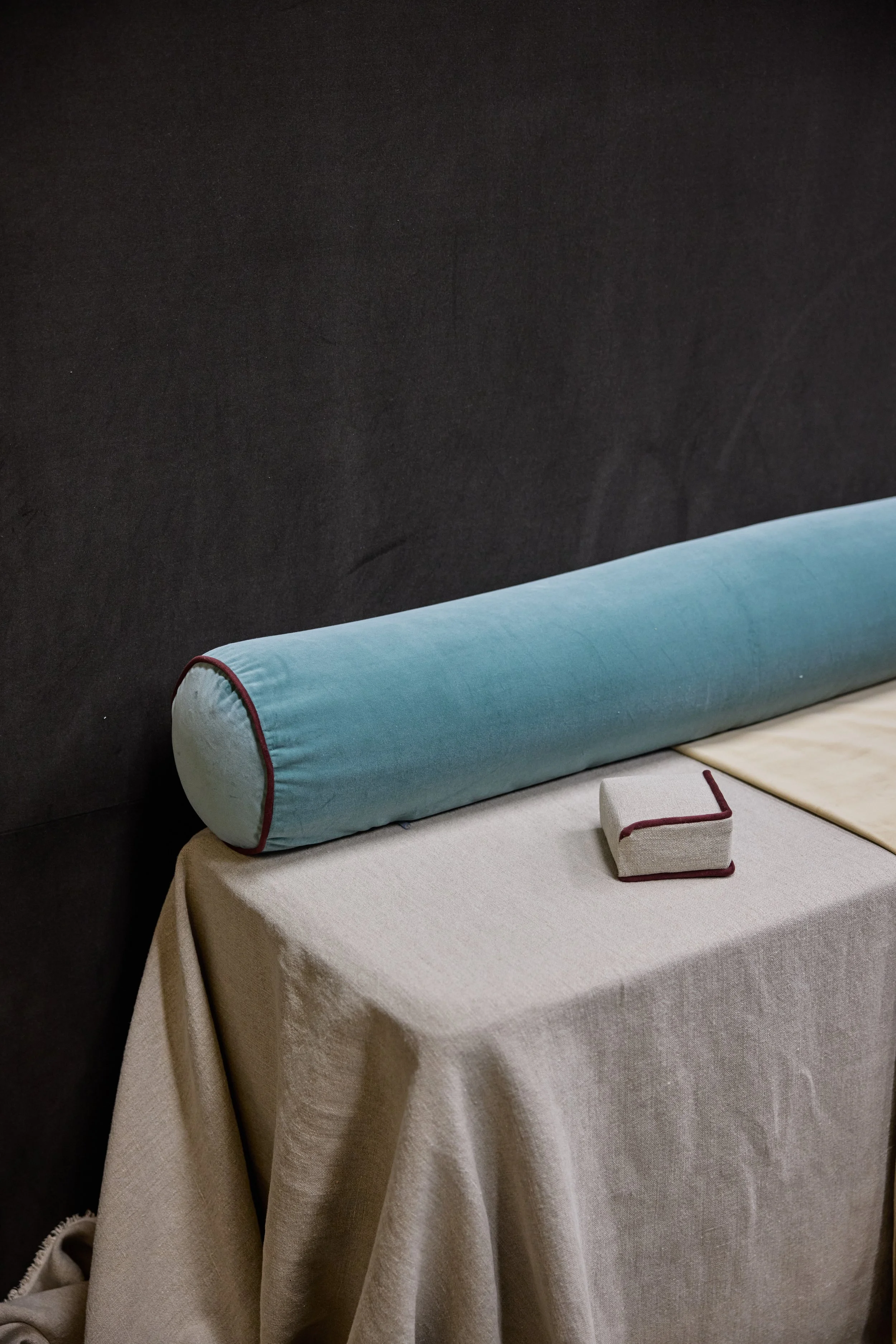 A round, light blue bolster pillow on a beige tablecloth-covered table, with a small L-shaped cushion in front of it, against a dark wall.