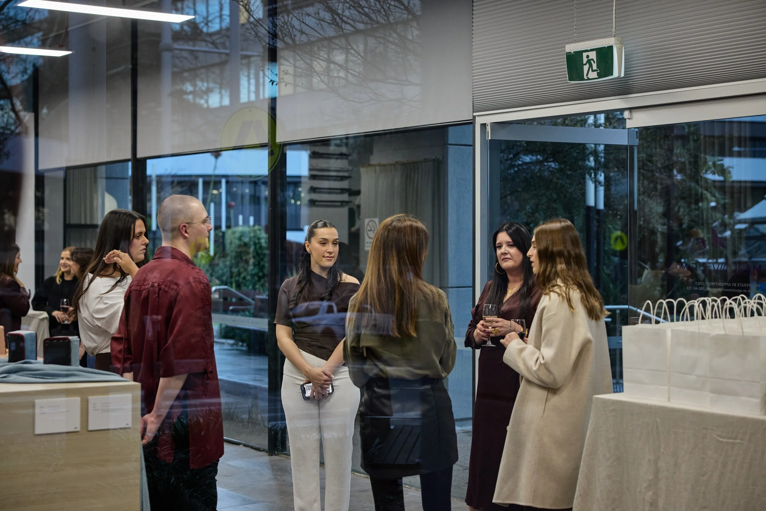 Group of people socializing at an indoor event near a glass entrance with an emergency exit sign above. Some are holding wine glasses.