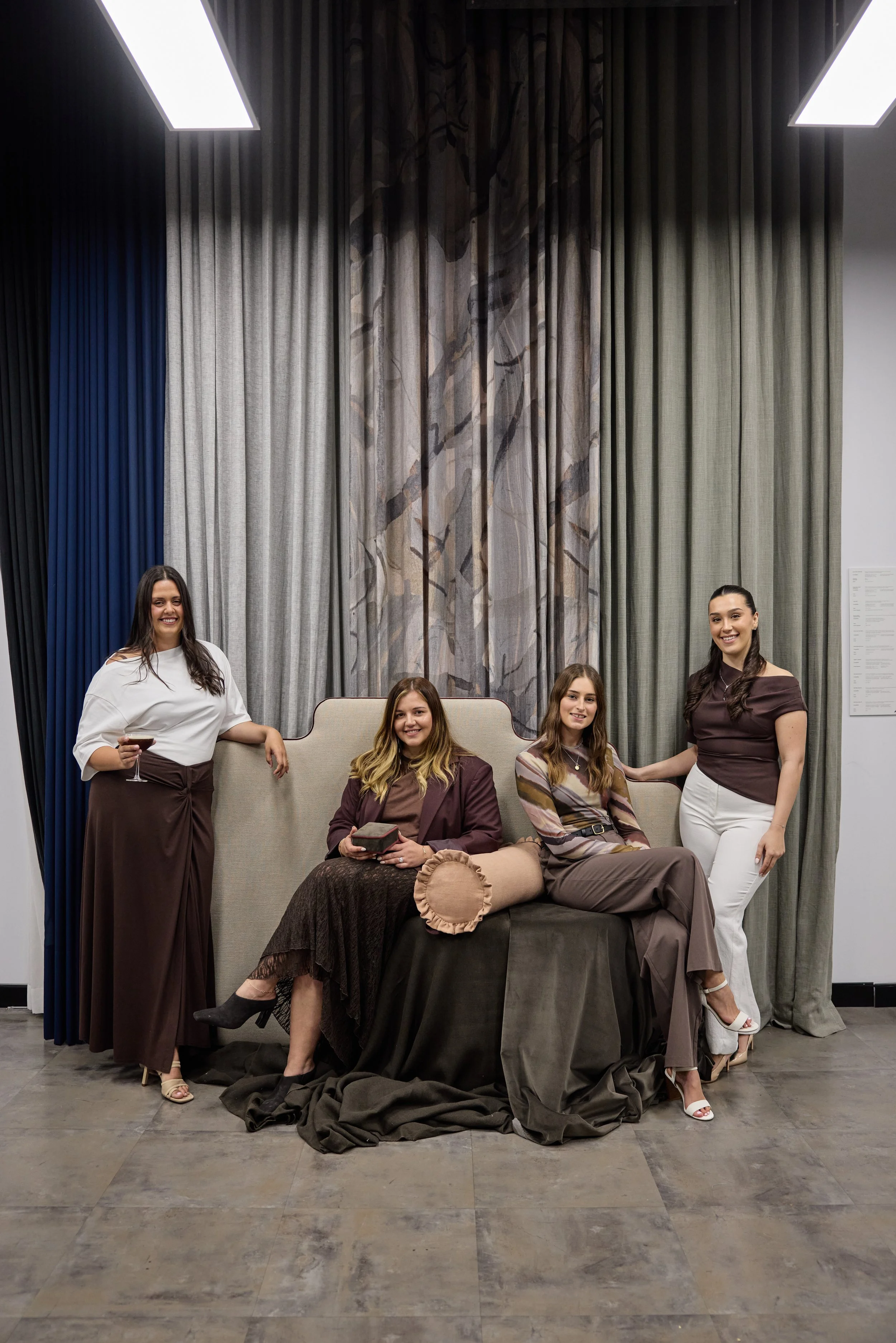 Four women in fashionable attire posing in front of a large, elaborate curtain backdrop, with two standing and two seated on a sofa.