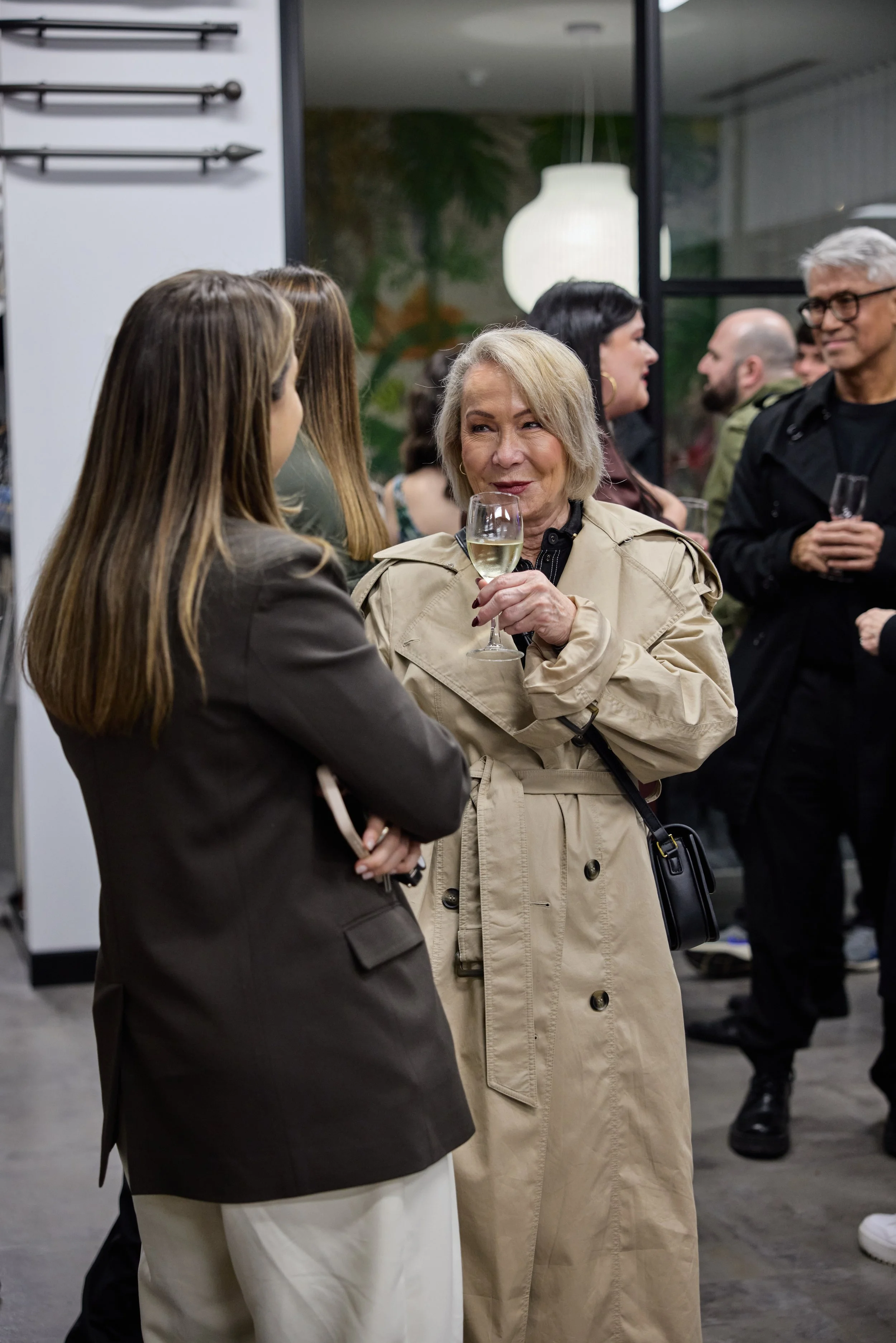 People socializing at an indoor event, with a woman in a beige trench coat holding a glass of white wine and talking to a woman in a dark blazer.
