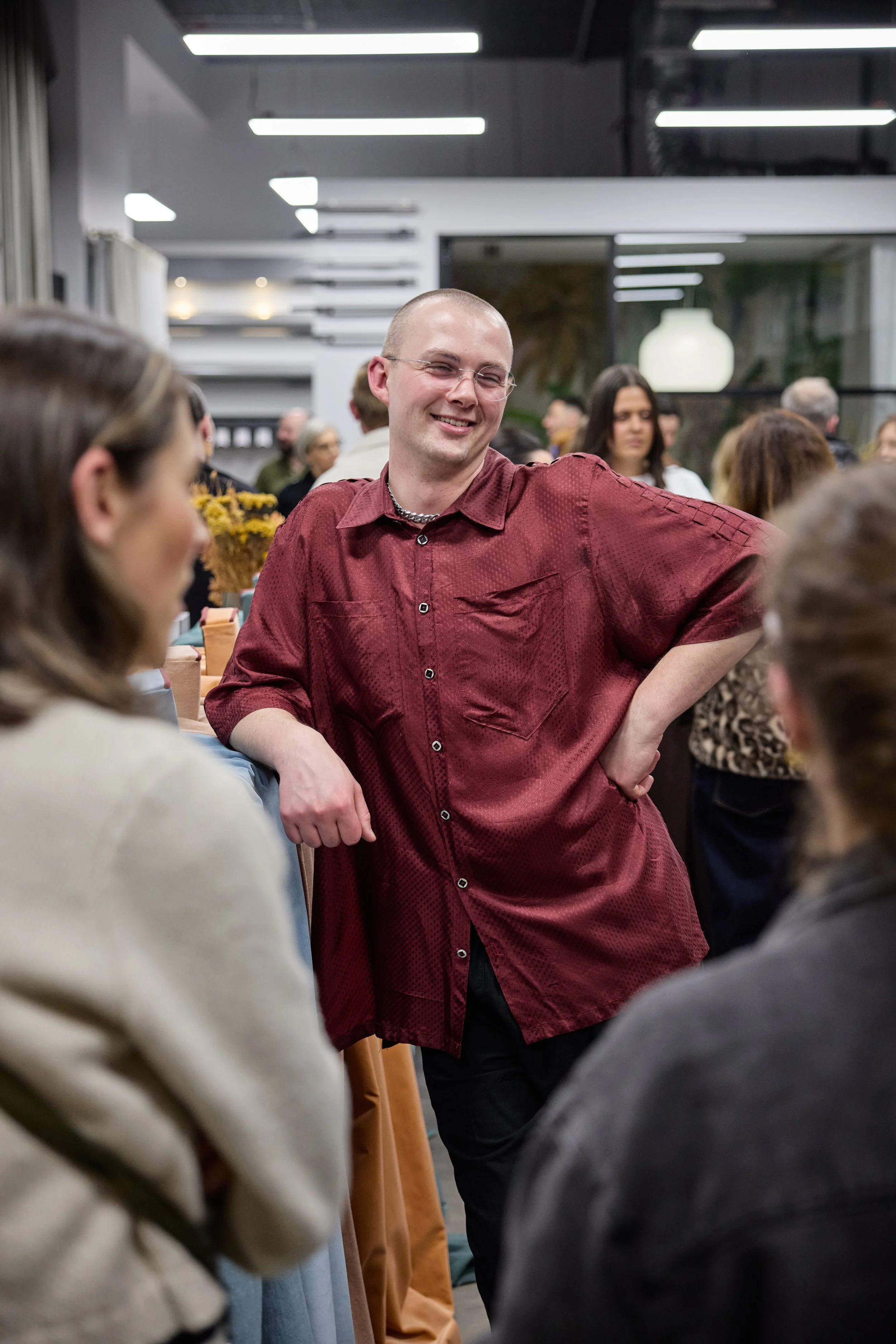 A man in a red shirt smiling and chatting with two women at an indoor social gathering.