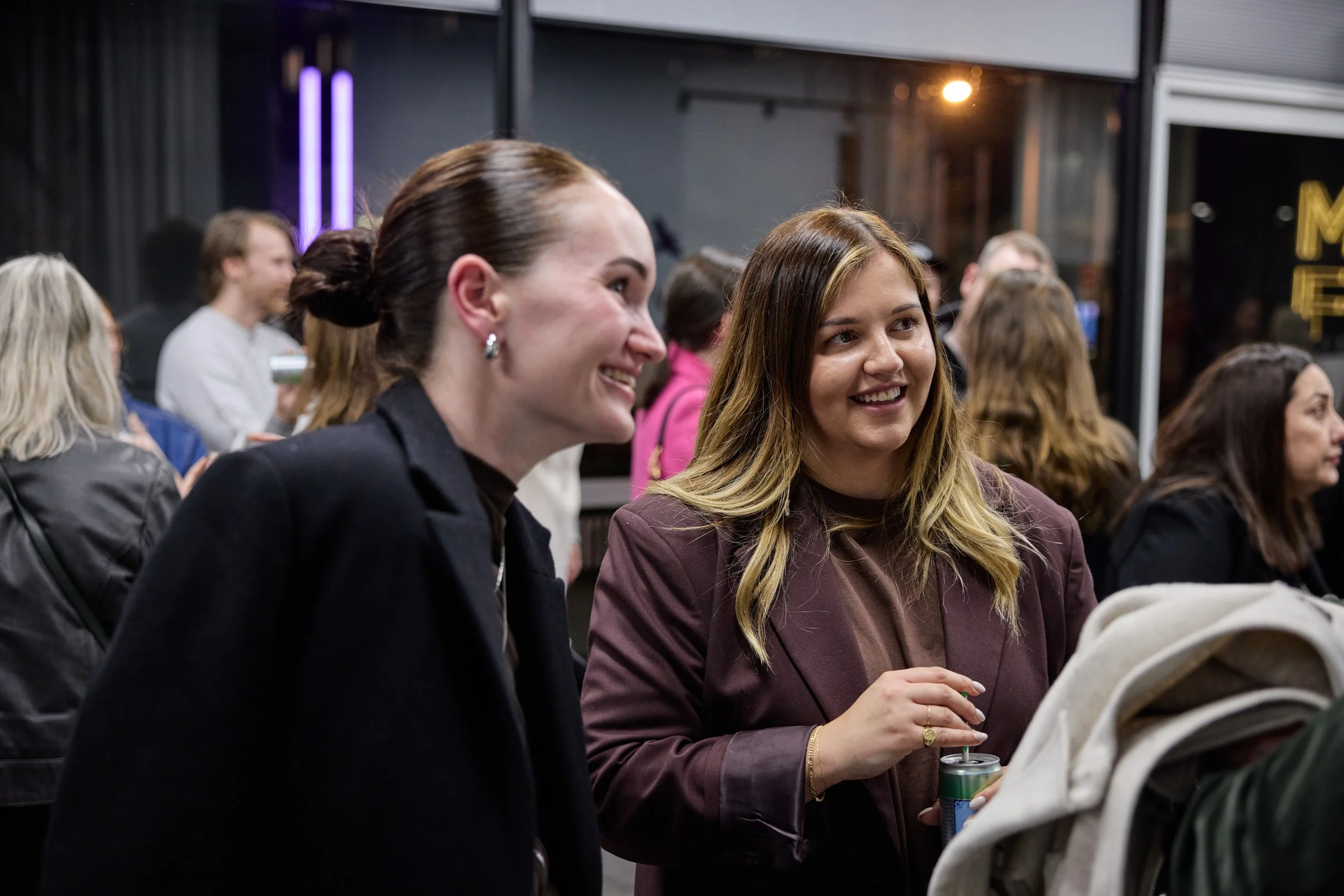 Two women smiling and talking at a social gathering at night, holding drinks, with others in the background at an indoor event.
