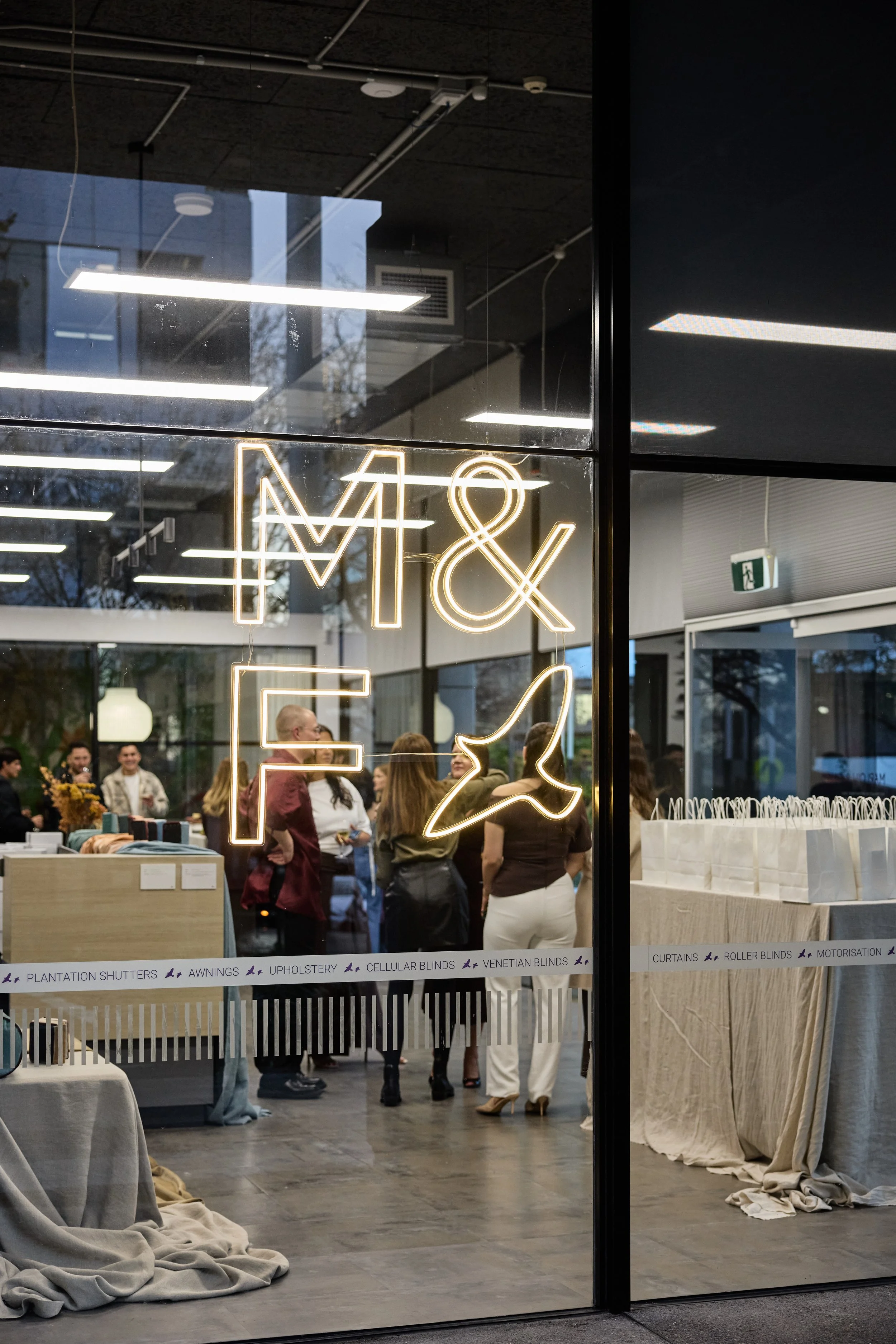 People gathered inside a store with a glass front, neon sign reading 'M & F', and various products on display.