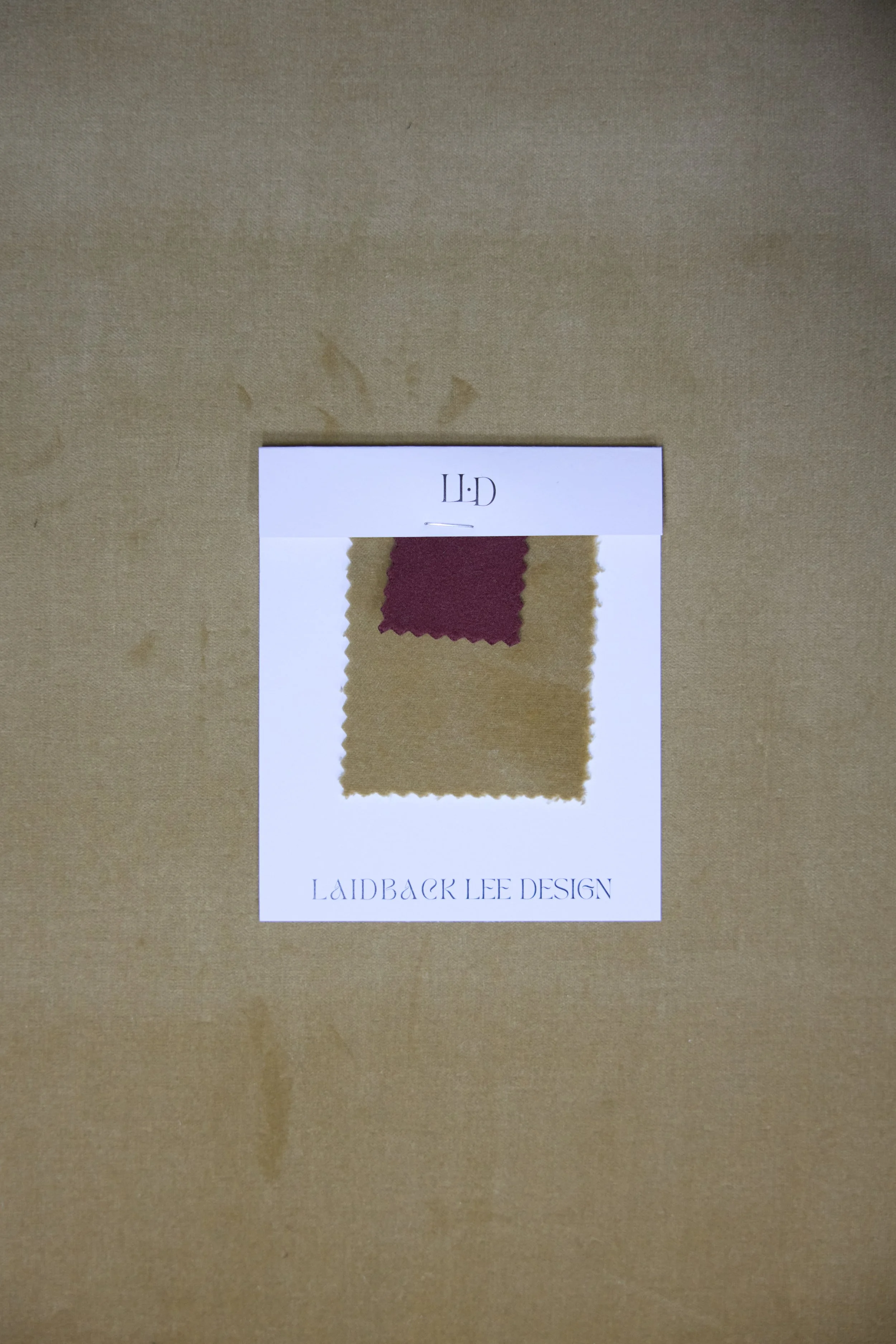 A fabric swatch sample card from LaidBack Lee Design with two fabric pieces, one maroon and one beige, attached to a white card, placed on a brown surface.
