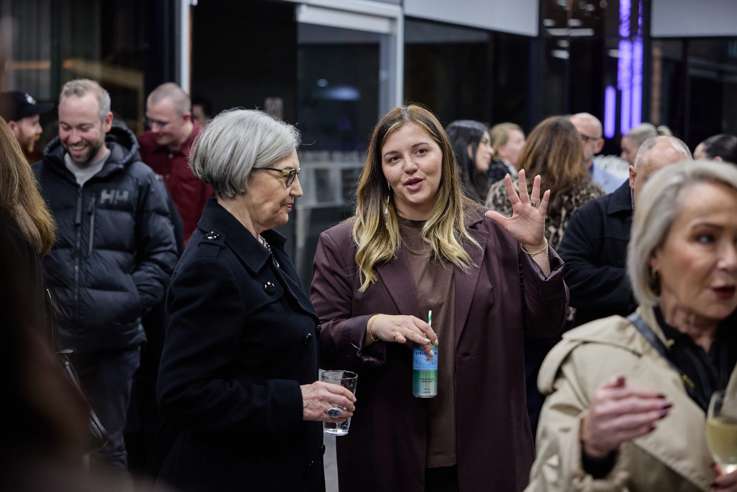 People socializing at an indoor gathering, including a young woman with long blonde hair wearing a brown jacket, speaking and gesturing with her hand, and an older woman with short gray hair and glasses holding a drink.