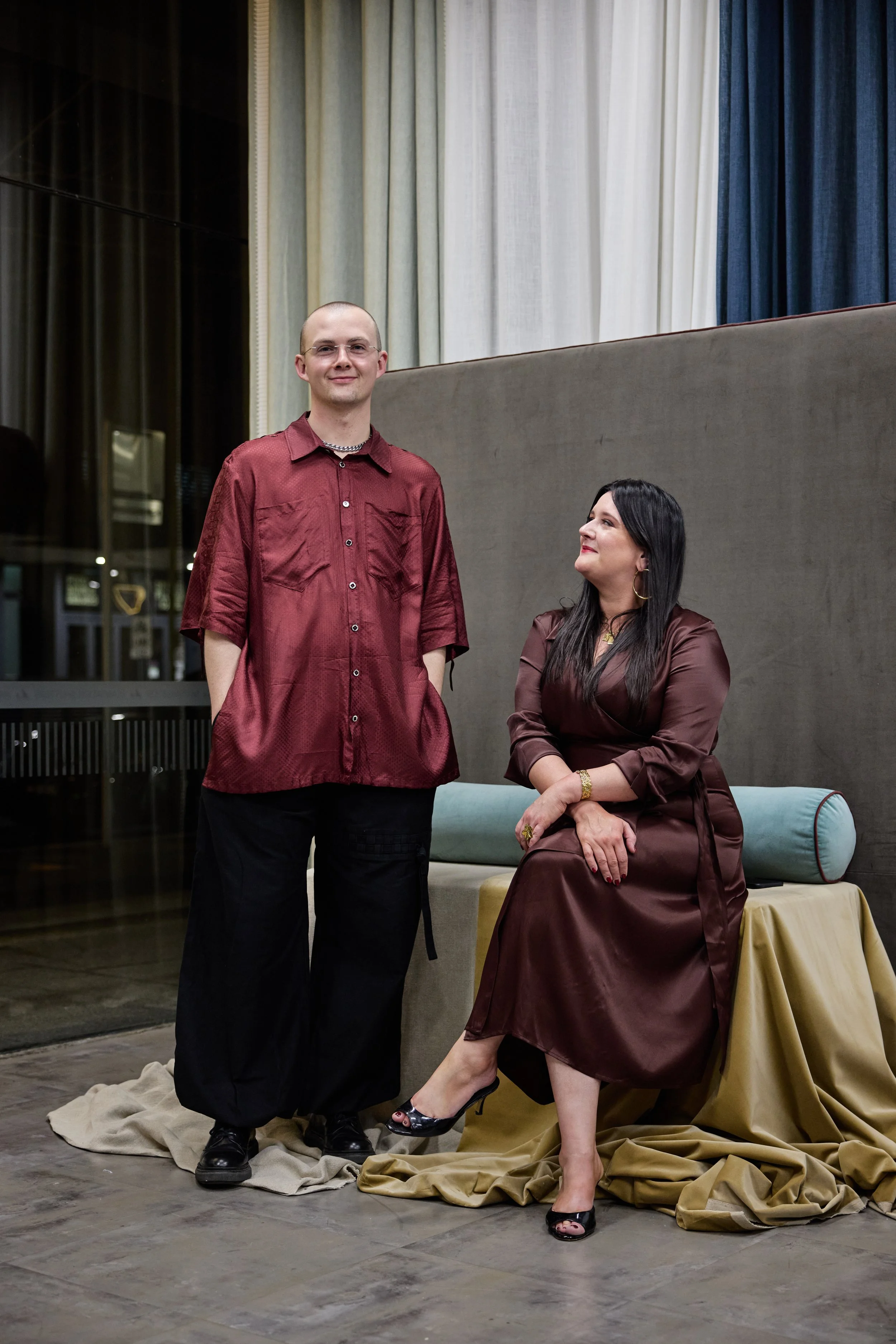 A man standing and a woman sitting on a bench inside a building with curtains and large glass windows.