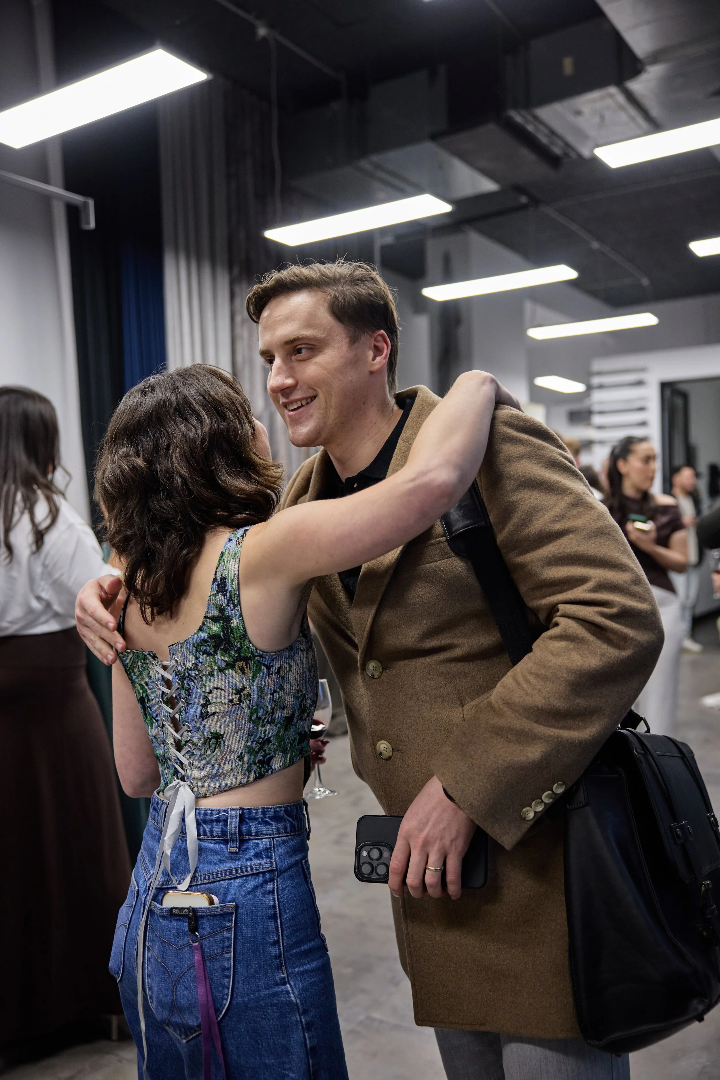 Two people, a man and a woman, hugging at an indoor social gathering or party. The woman is wearing a sleeveless floral top and jeans, and the man is wearing a tan coat with a black bag and carrying a smartphone. They are smiling and appear to be hap