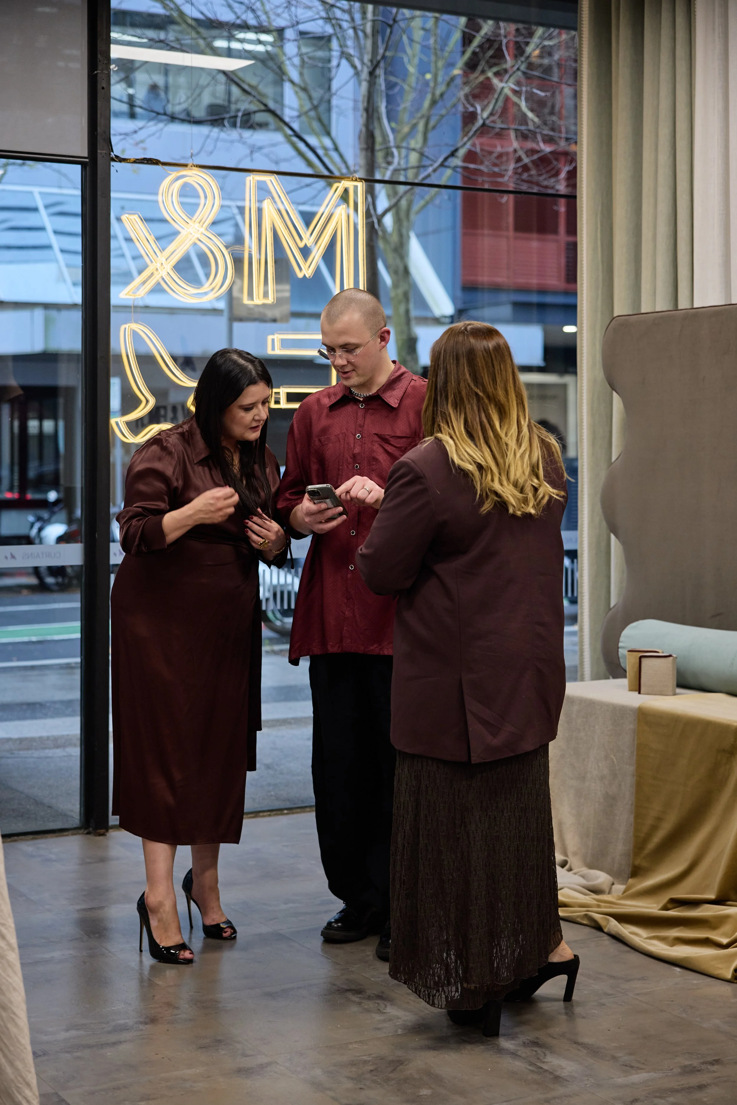 Three people indoors wearing dark red and maroon clothing, standing in front of a glass window with neon sign, looking at a smartphone.