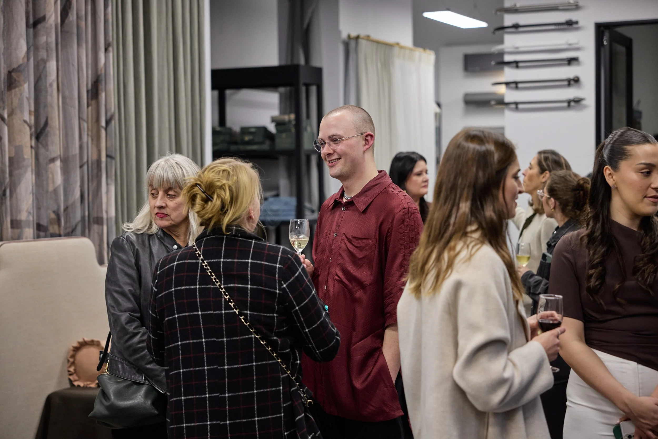 People socializing at a gathering, holding glasses of wine, in a room with shelves and curtains.