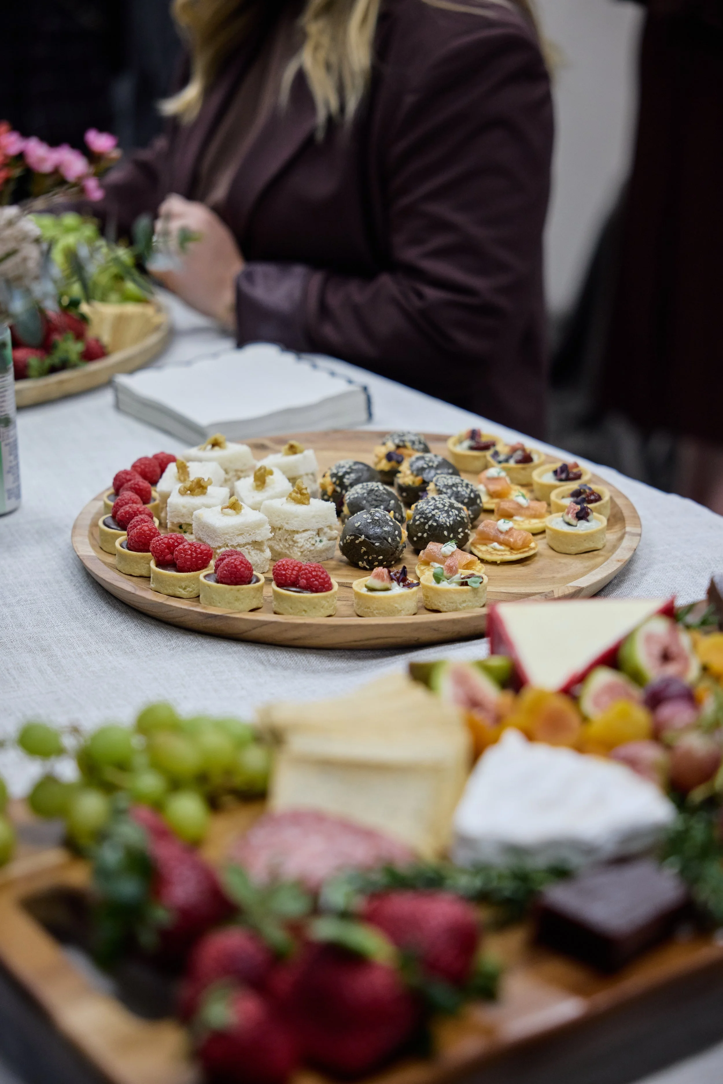 assorted small desserts like tarts and cakes on a wooden platter, raspberries and other fruit decorations, with a person in a dark jacket sitting at a table with more food and floral decorations in the background.