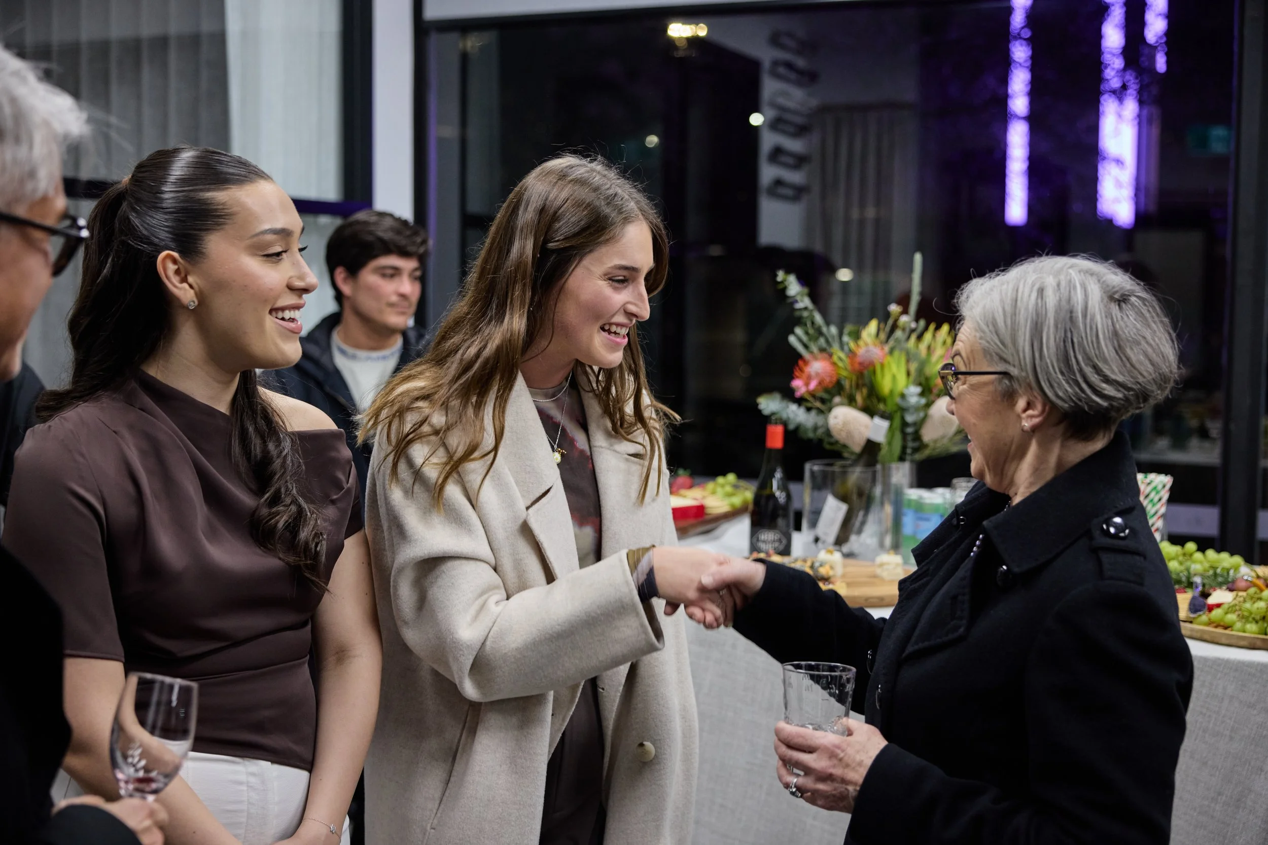 A group of people at a social gathering, with a woman in a beige coat shaking hands with an older woman with gray hair and glasses, in front of a table with flowers, wine, and snacks.