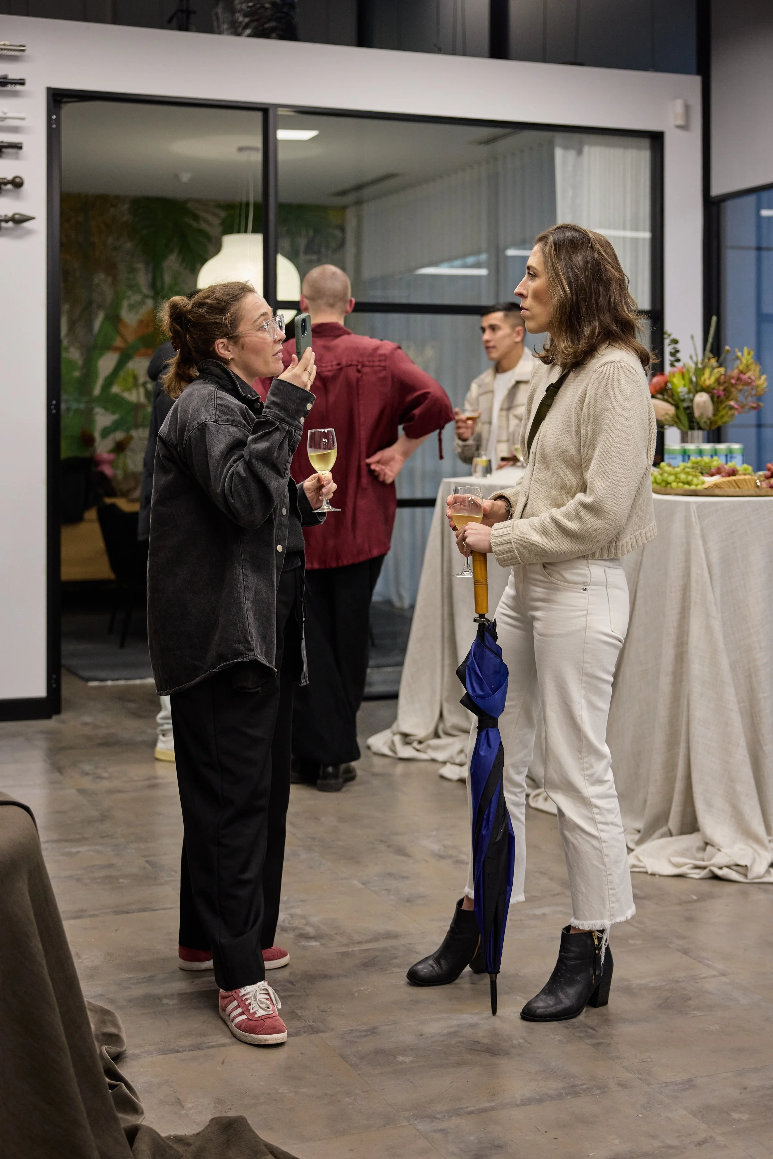 Two women are having a conversation at a social gathering, each holding a glass of white wine, with one woman taking a photo or video of the other.