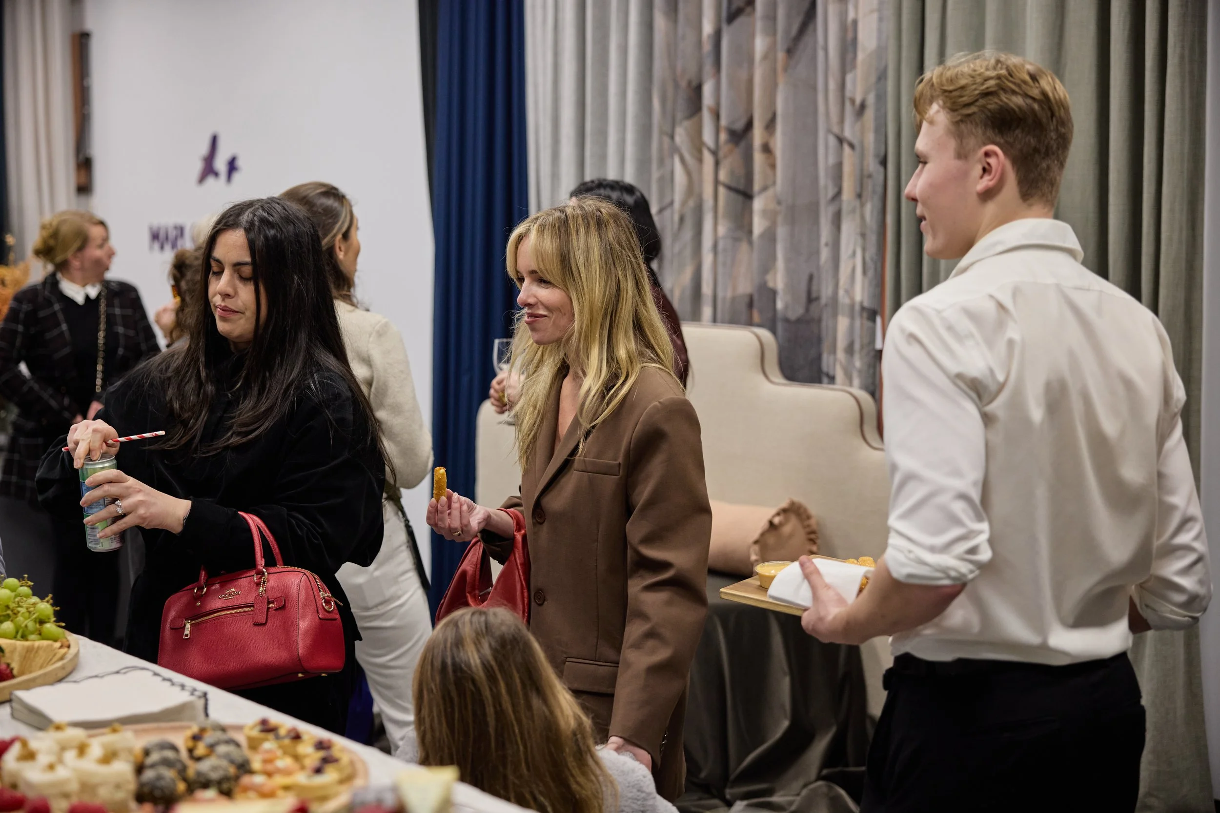 Group of people socializing at a party with food and drinks, including women and a man holding a tray of snacks, in a room with curtains and a decorated wall.