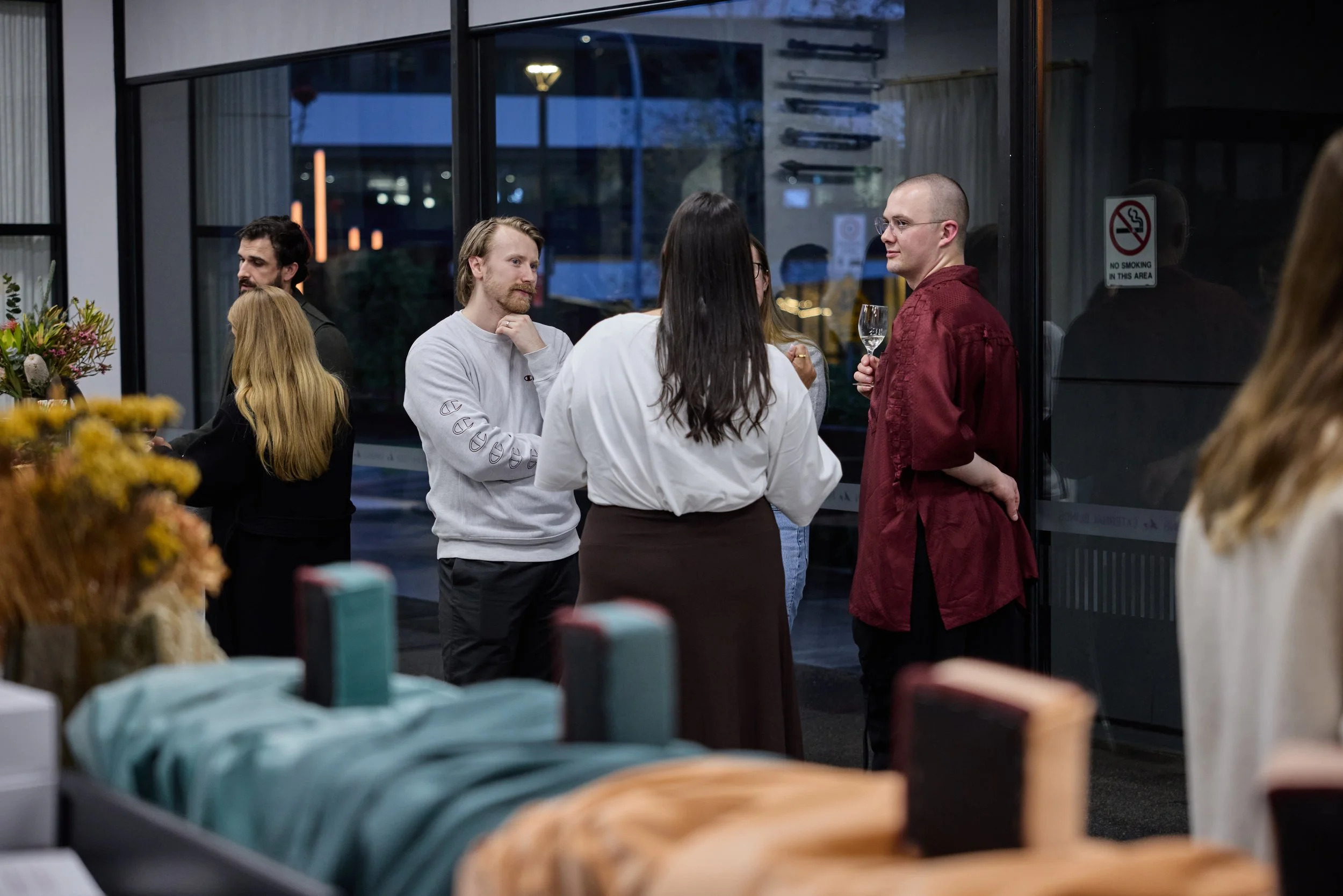 People socializing at a gathering inside a venue near large glass windows in the evening or night.